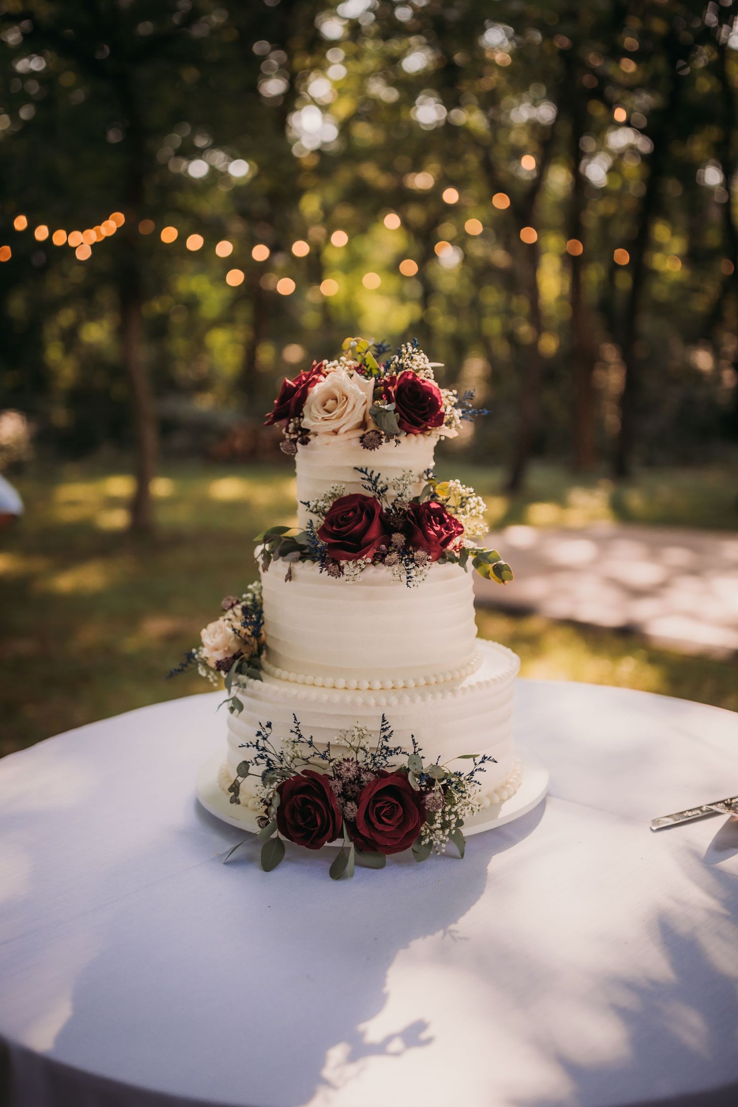Three-tiered white wedding cake adorned with deep red roses and greenery under string lights.