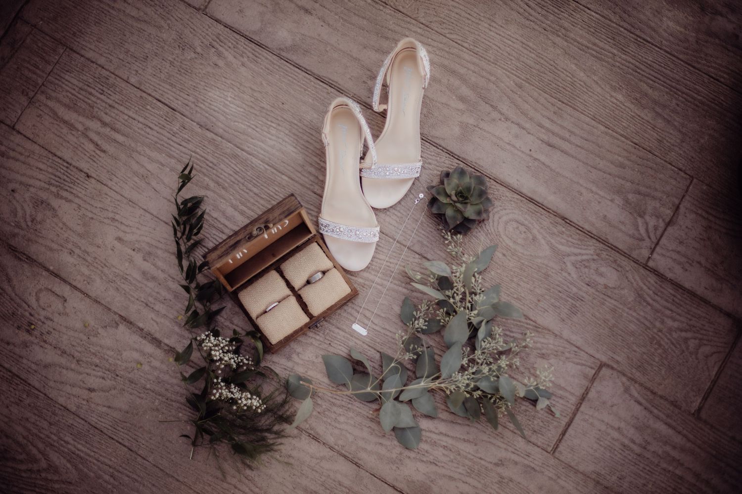 Wedding shoes, wooden ring box and eucalyptus branches styled on wooden floor.