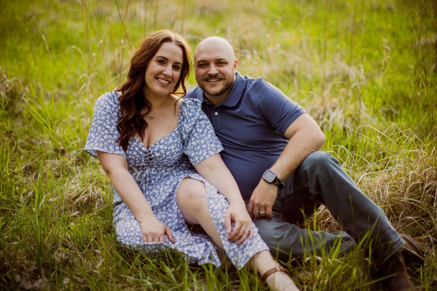 A loving couple sits together in tall grass during a romantic outdoor photoshoot.