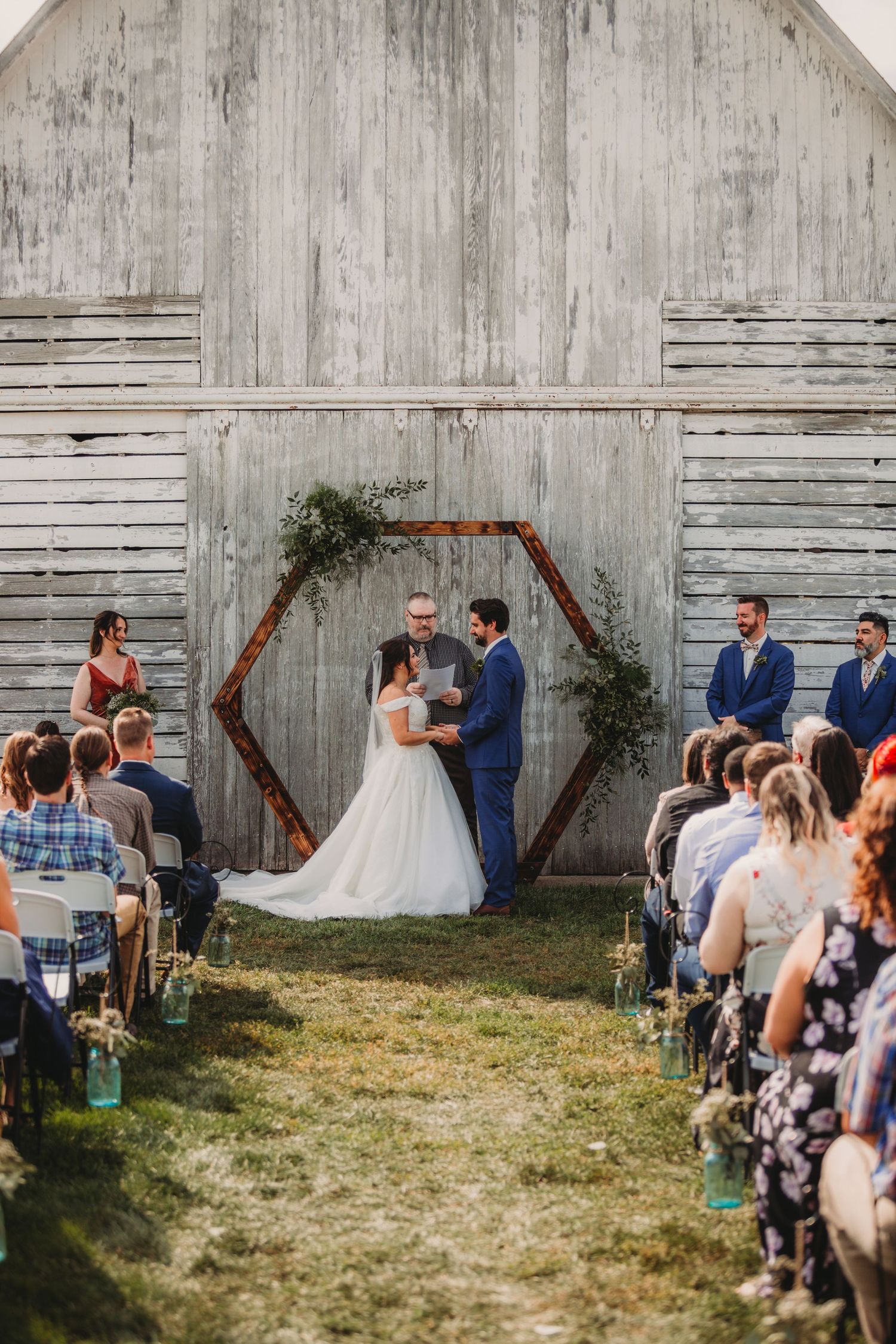 An outdoor wedding ceremony takes place in front of a rustic barn wall with a wooden triangle arch and gathered guests.
