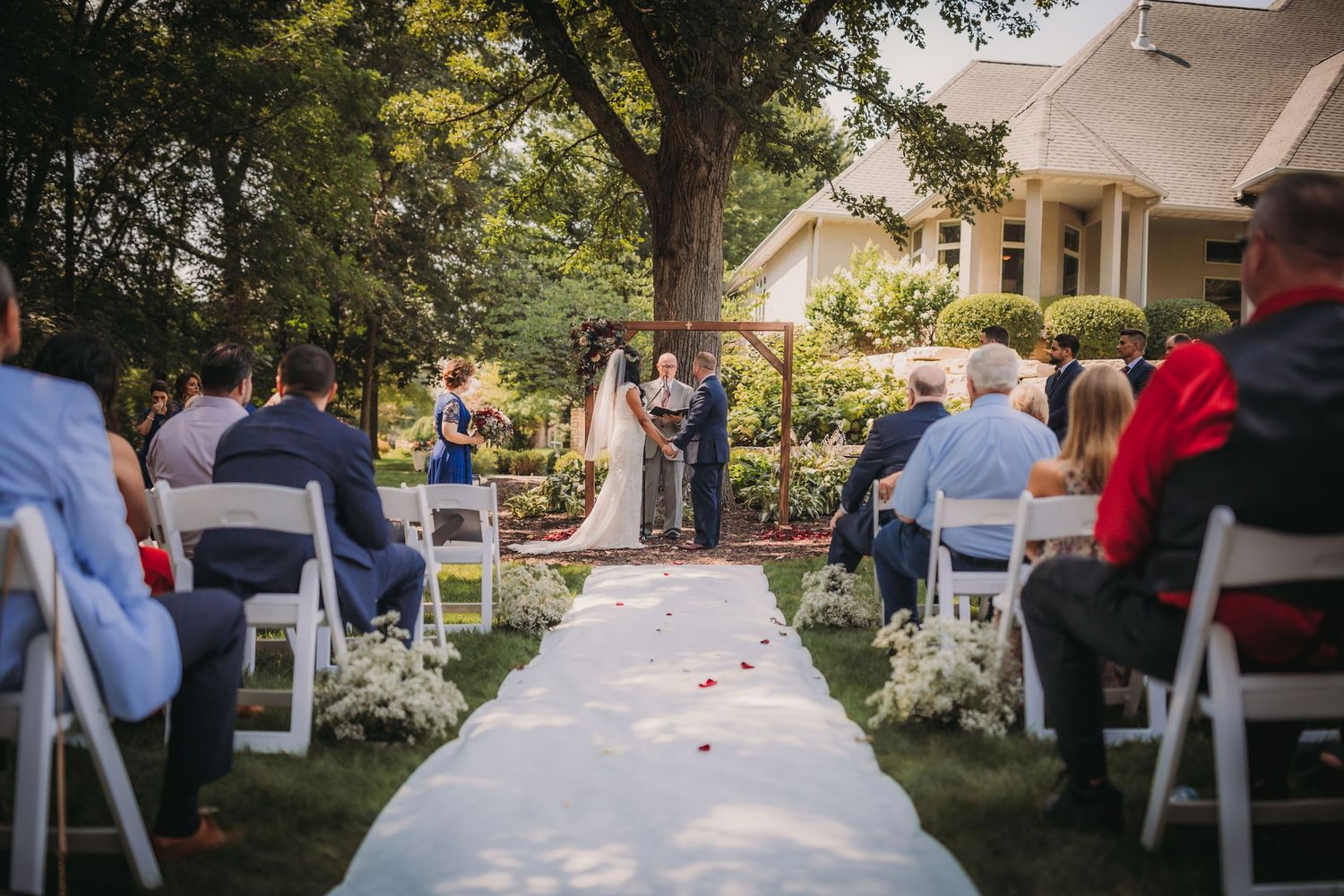 Garden wedding ceremony setup features white chairs and floral arrangements beneath shady trees.