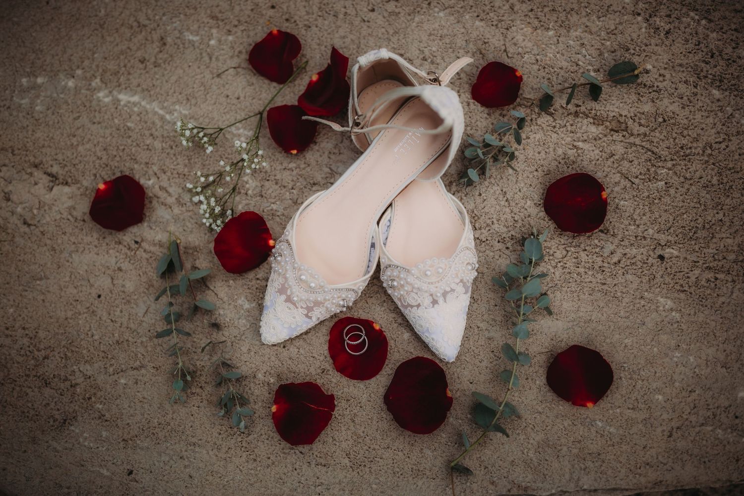 White bridal shoes arranged with deep red roses and eucalyptus on concrete surface.