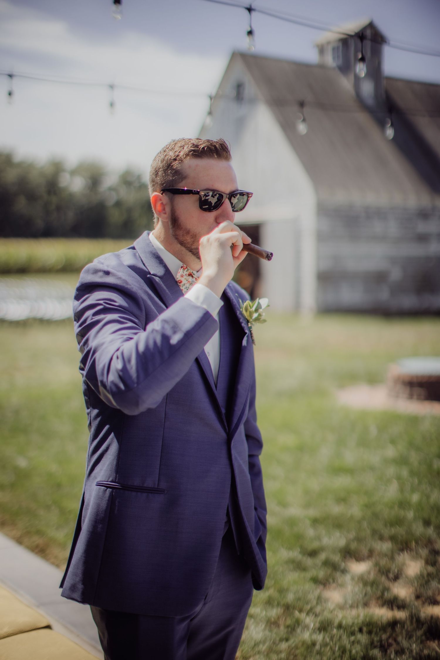 Groomsman in navy suit and sunglasses enjoying a cigar with white barn in background.