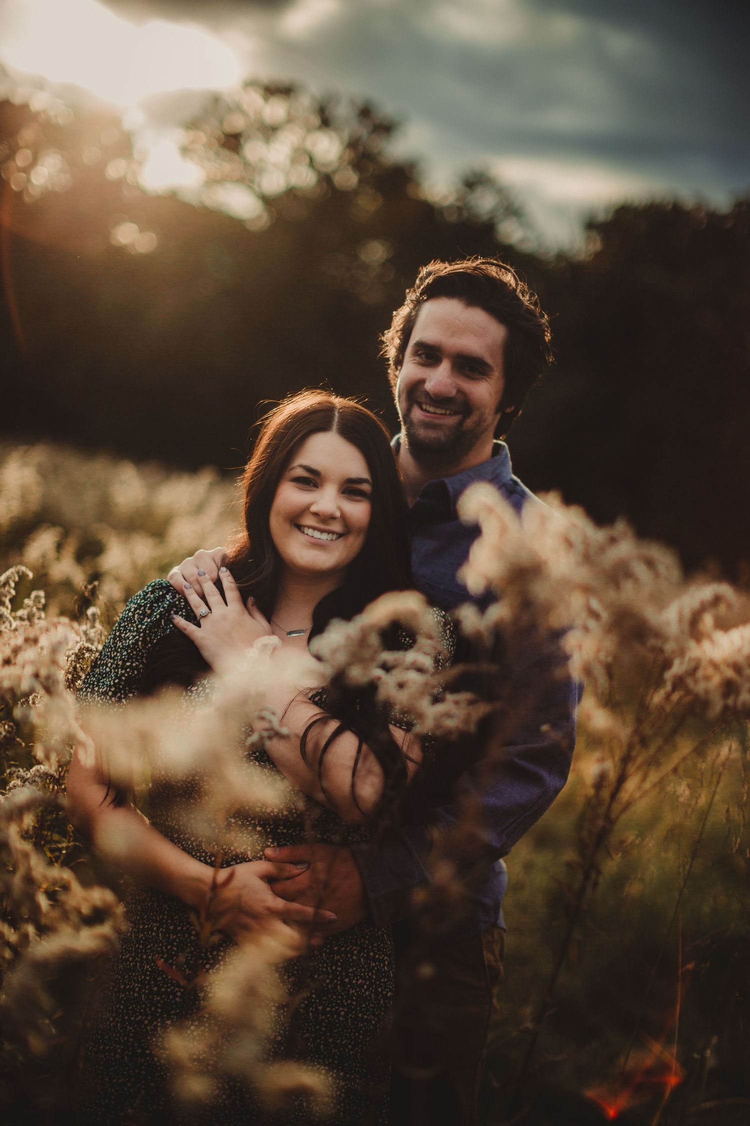Couple cuddling with their pet dog in golden sunset field.