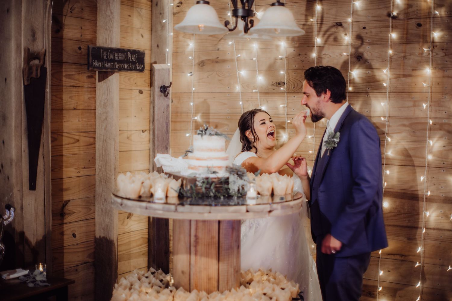 Couple shares a playful moment while cutting their wedding cake in a rustic venue with string lights.