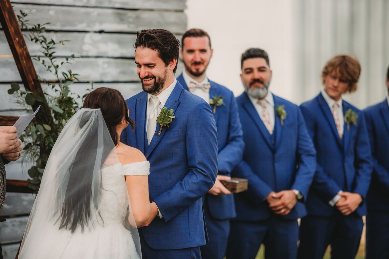 Wedding party members stand in formation wearing coordinated blue suits at outdoor venue.