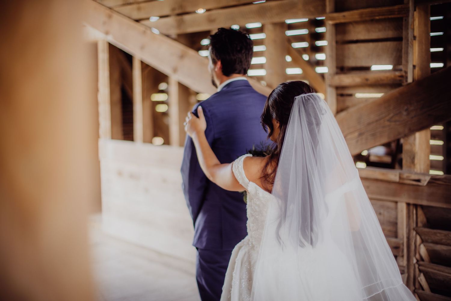 A bride in a white gown approaches her groom during their first look in a wooden barn.