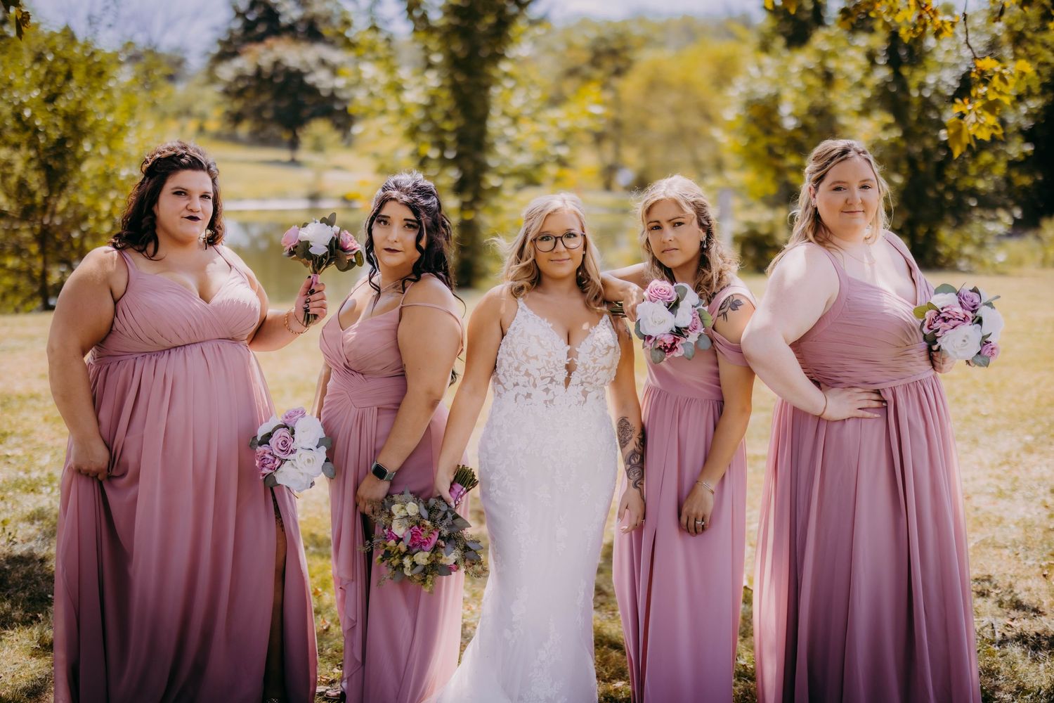 Wedding party stands together outdoors in mauve bridesmaid dresses holding floral bouquets.