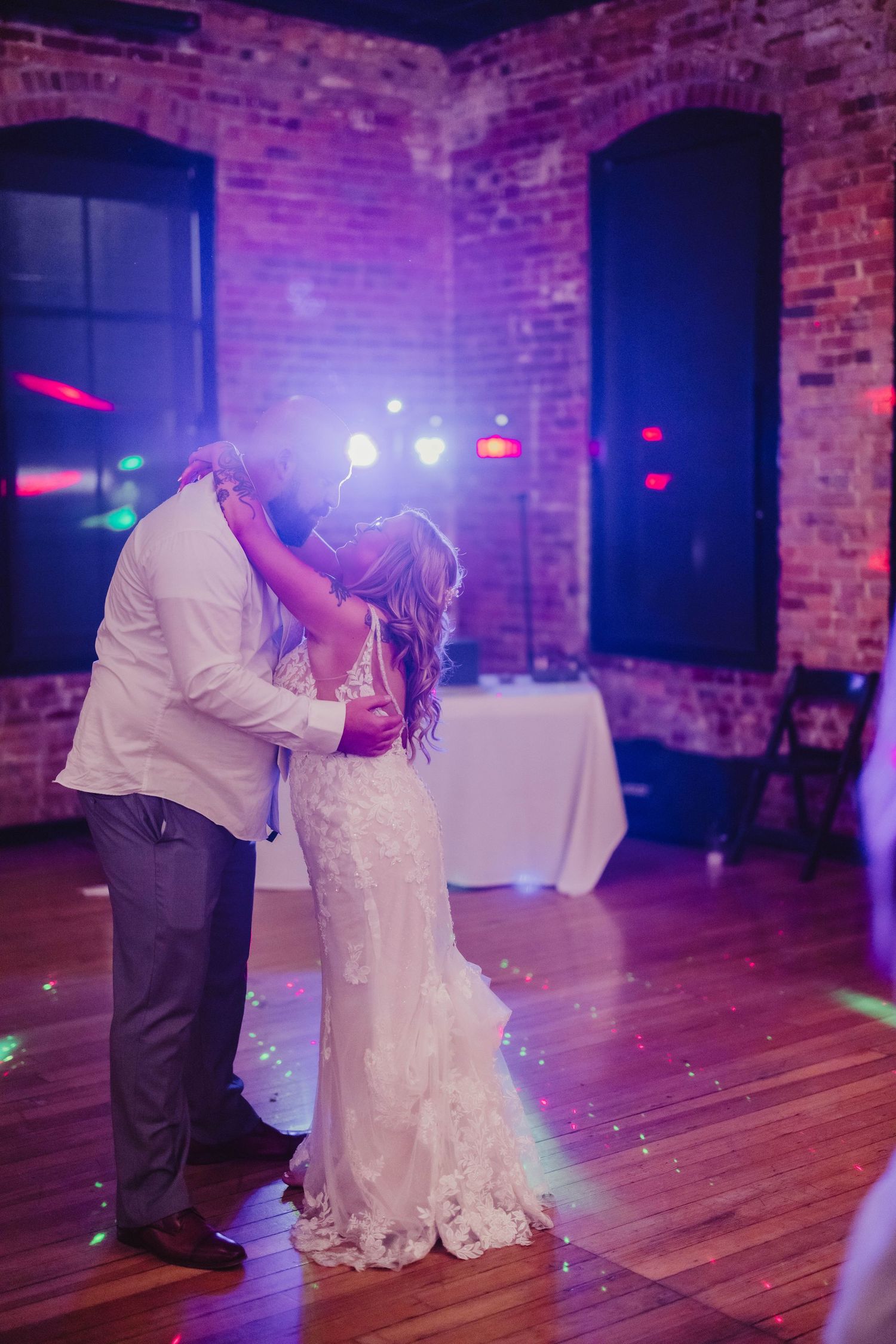 Couple dances in a purple-lit reception venue with exposed brick walls and hardwood floors.