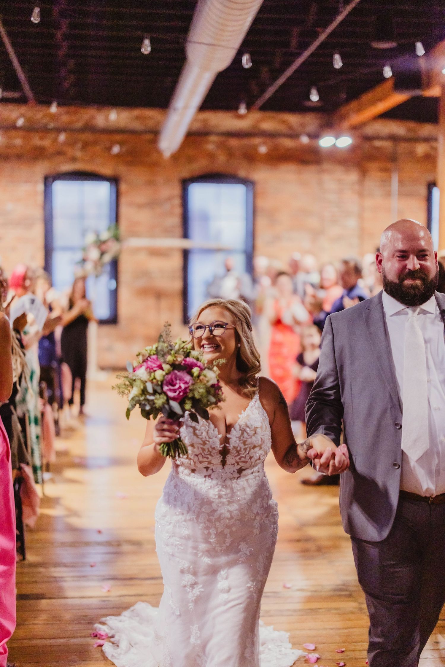 Wedding couple walks down aisle while guests celebrate in rustic brick venue with industrial details.