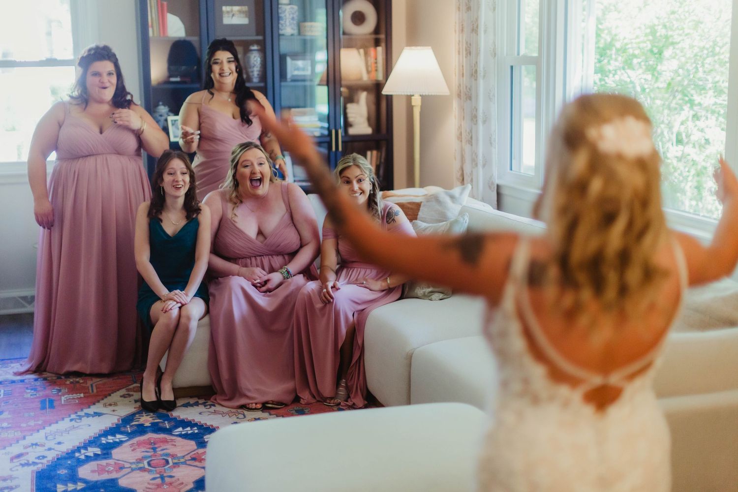 Bridesmaids in dusty rose dresses gather on a white couch during wedding preparations.