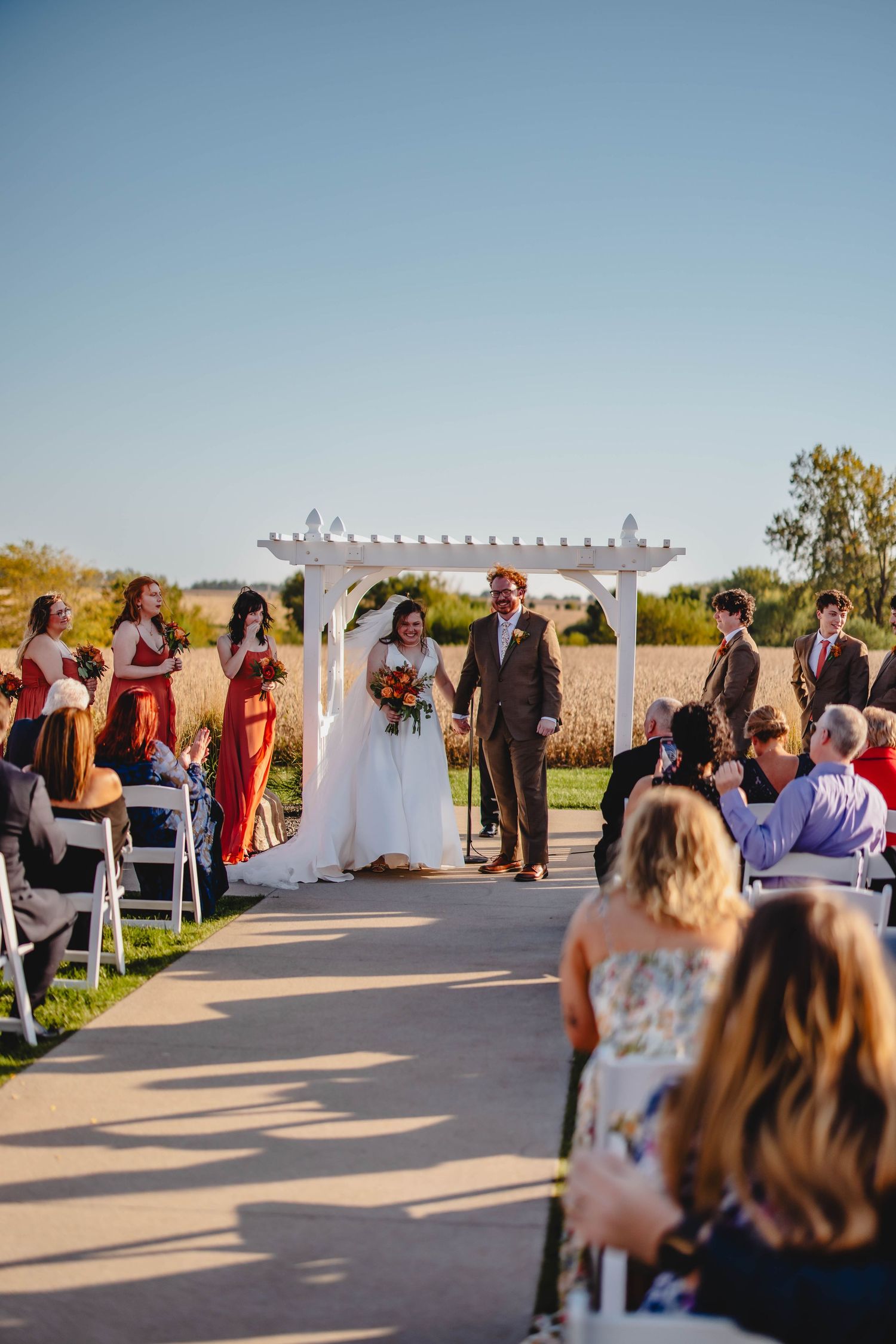 Wedding ceremony under a white pergola with bridesmaids in rust-colored dresses and guests seated on white chairs.