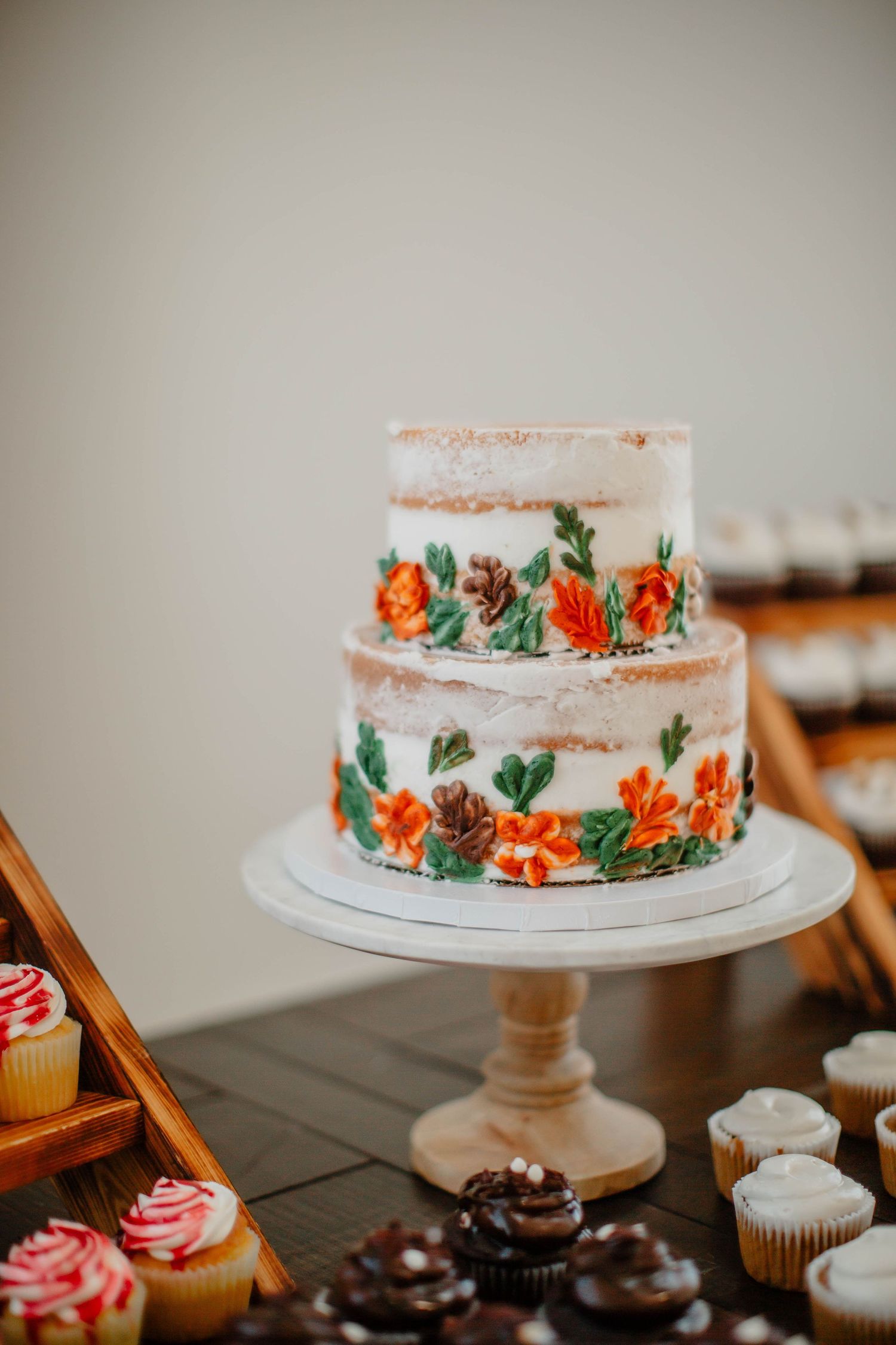 Semi-naked wedding cake with orange floral decorations displayed alongside cupcakes on a dessert table.