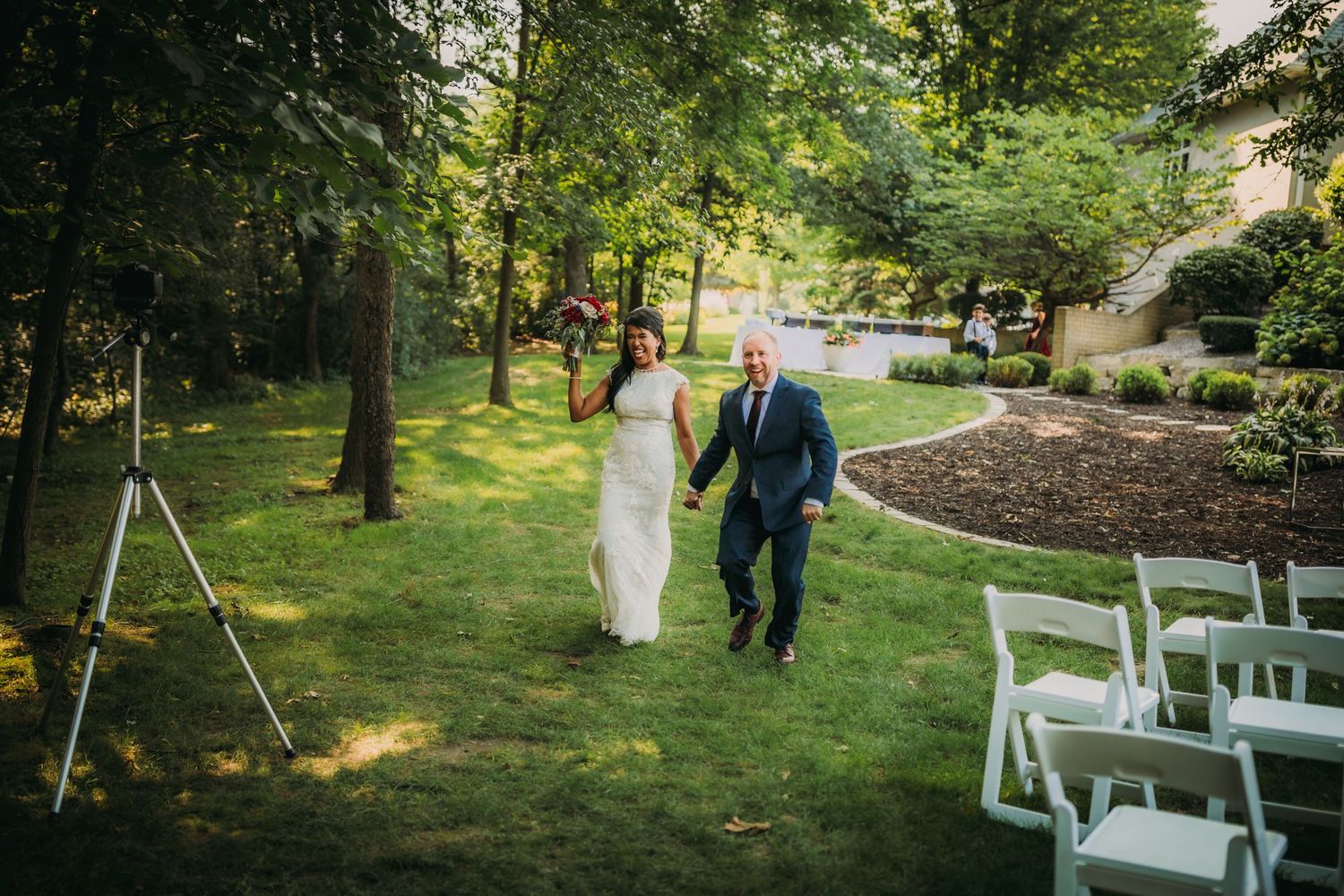 Wedding guests gather on a lush green lawn surrounded by tall trees.