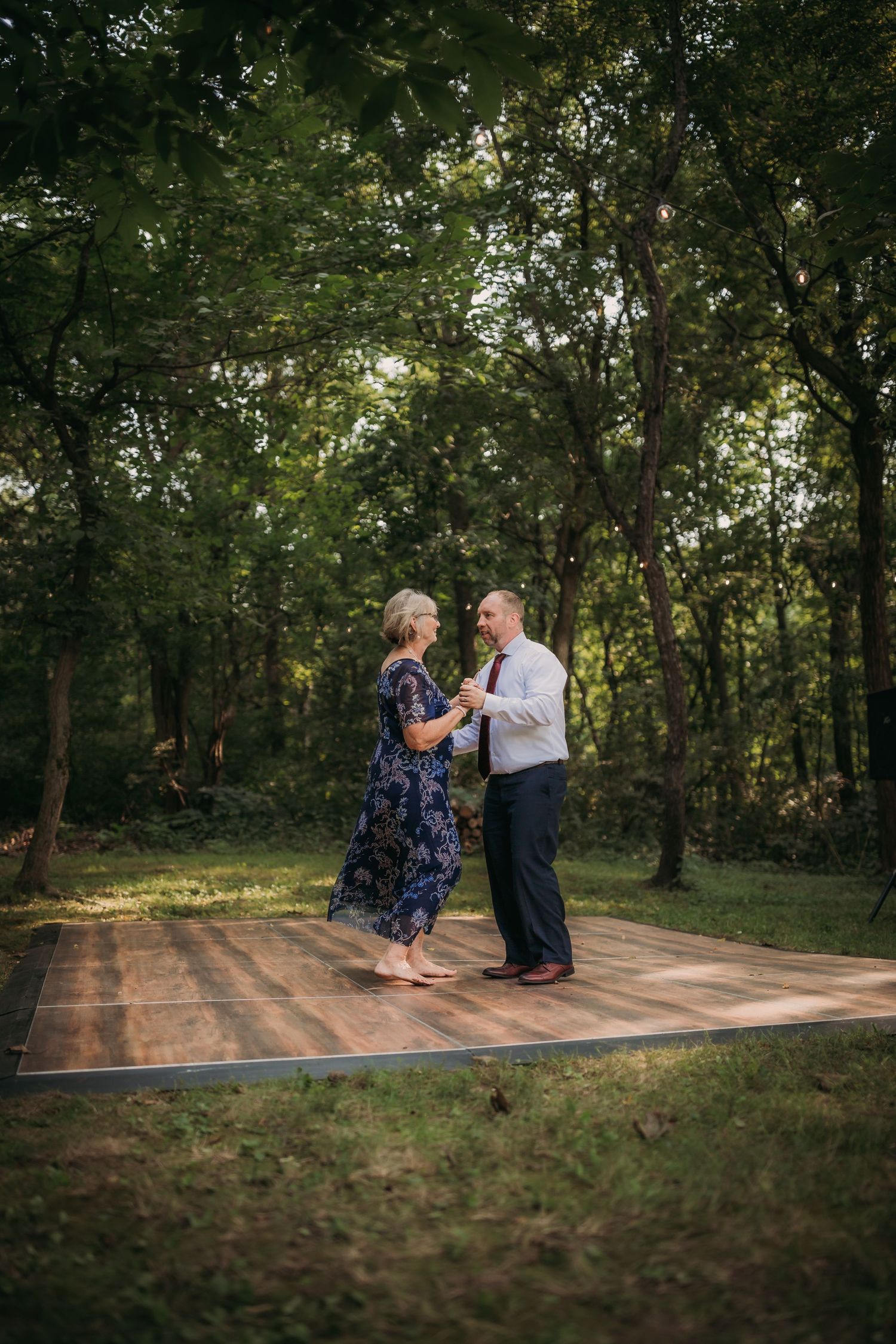 Couple dancing together on a wooden platform surrounded by trees and natural scenery at an outdoor event.
