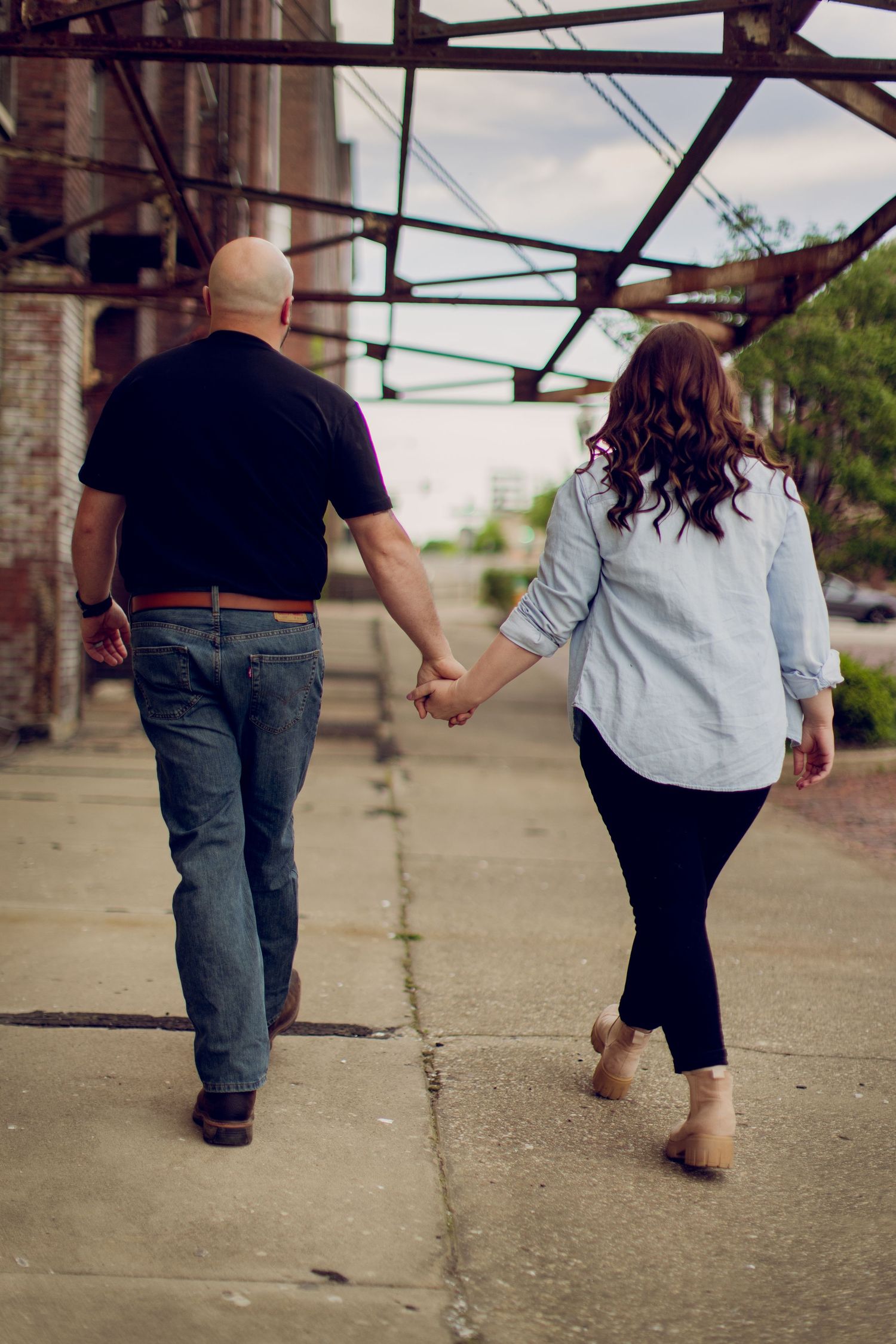 A couple takes a romantic walk holding hands beneath an urban overhead structure.