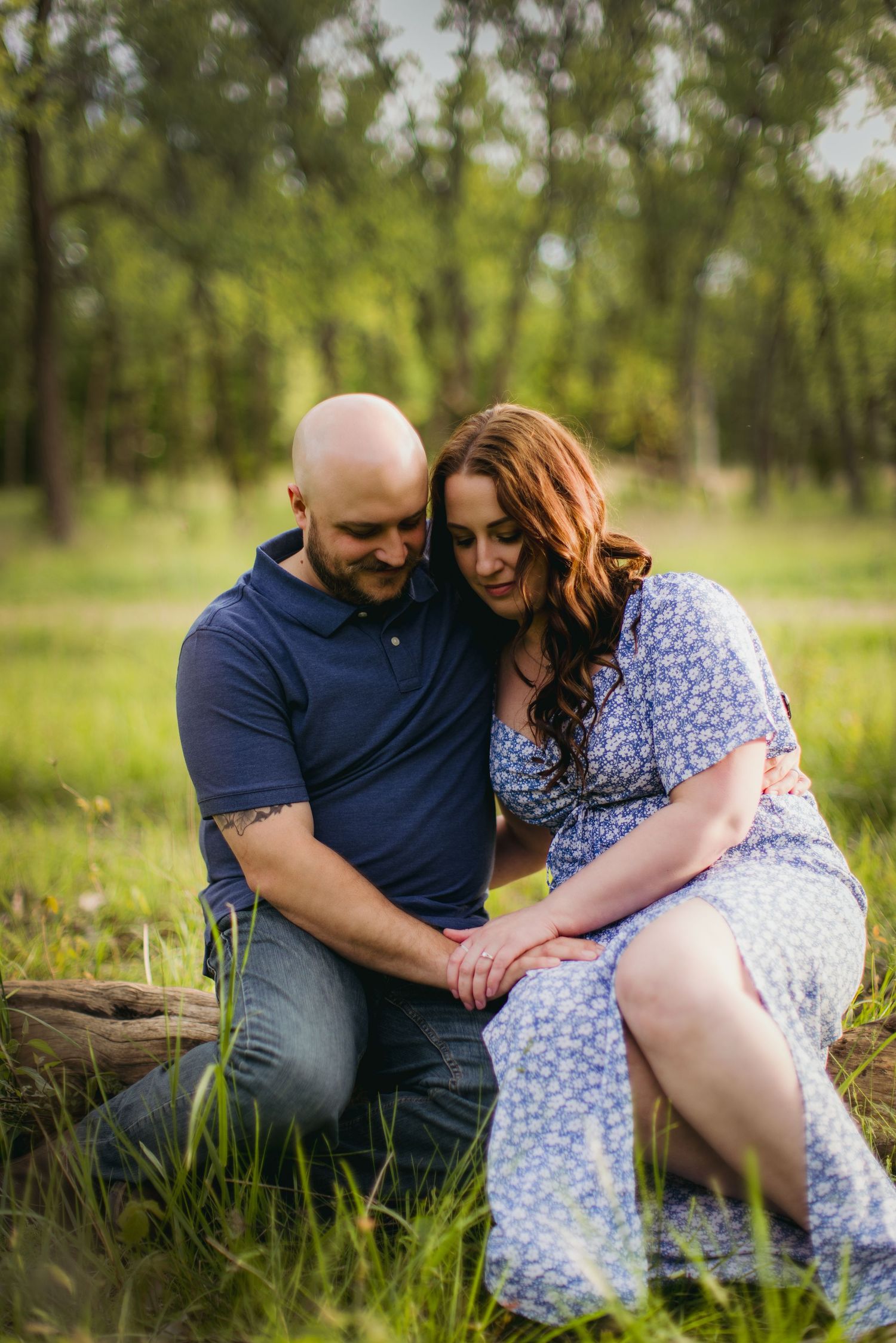 A couple in coordinating blue outfits shares a tender moment while sitting on a log in a sunlit forest.