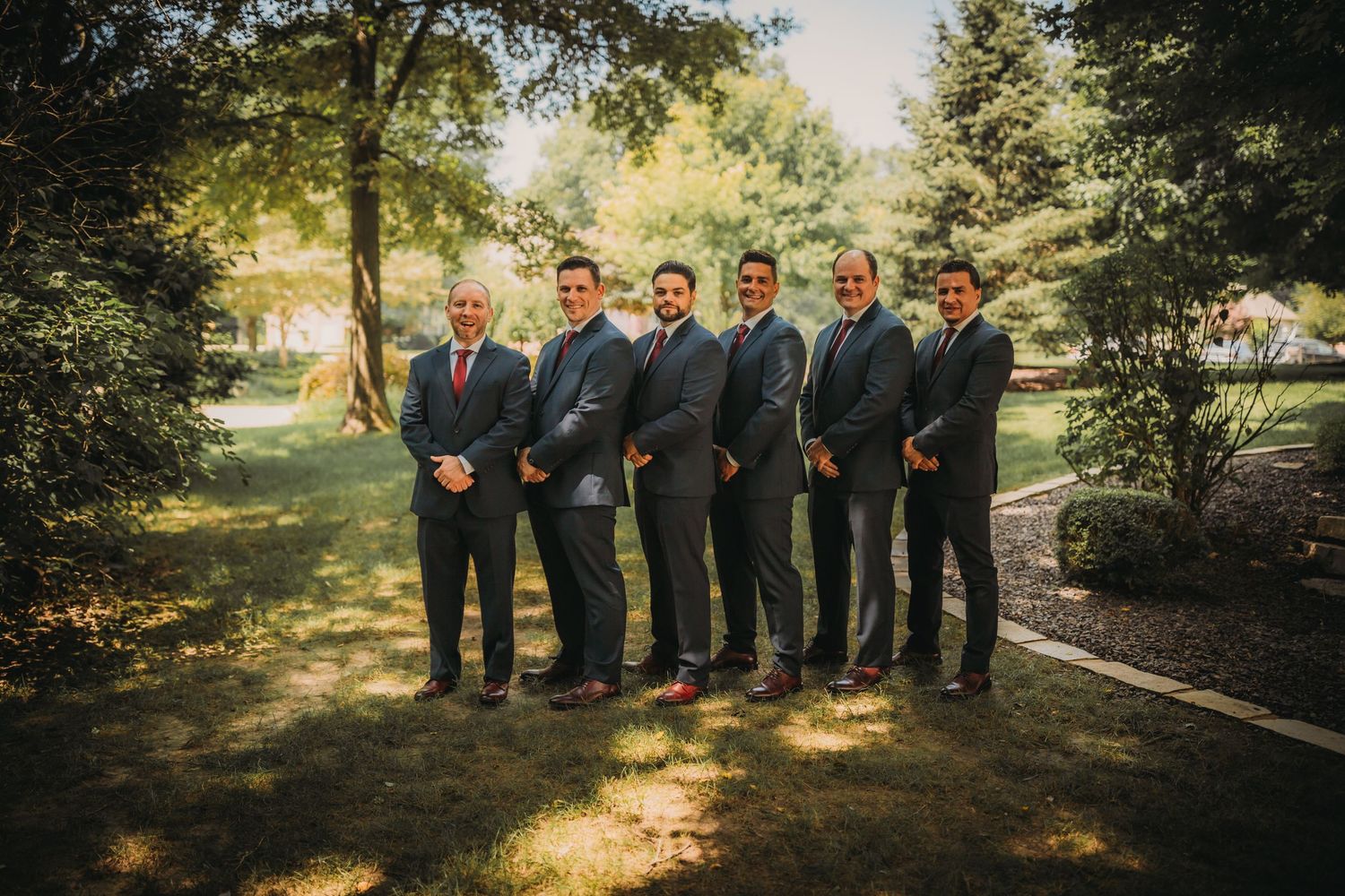 Groomsmen lined up in formal attire for wedding photos in a sunlit garden setting.