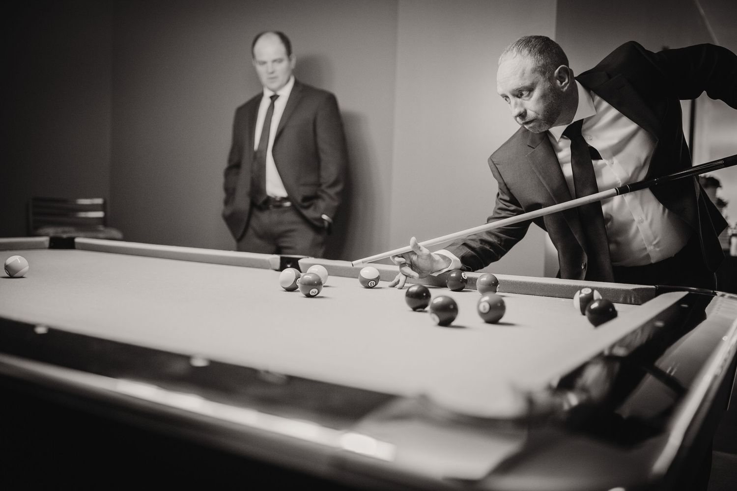 Two people play billiards at a pool table with one leaning over to take a shot in black and white.
