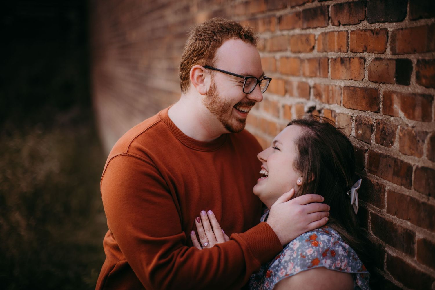 Candid moment shared between a couple posed in front of a weathered brick wall.