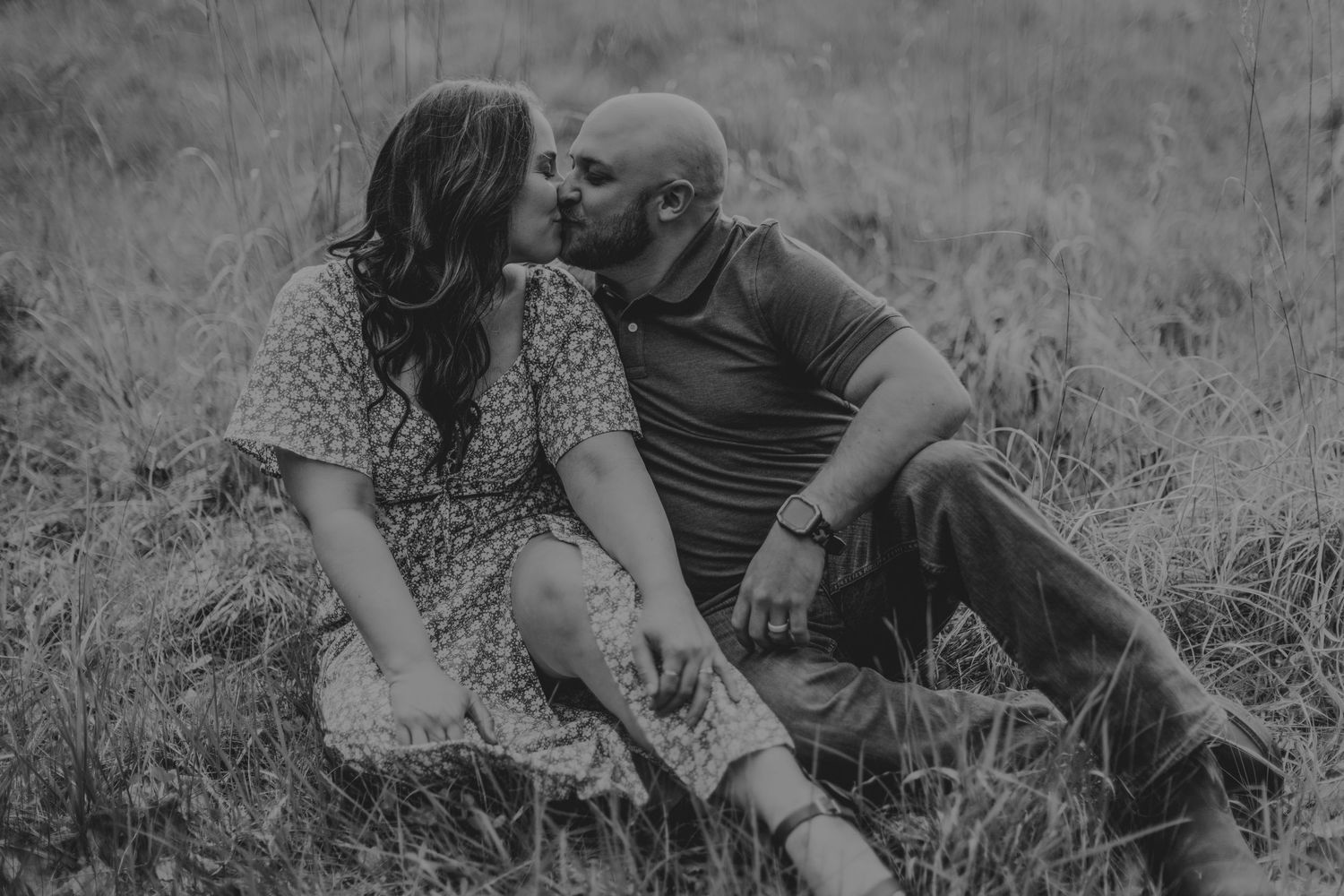 An intimate black and white portrait of a couple sitting close in a field of grass.