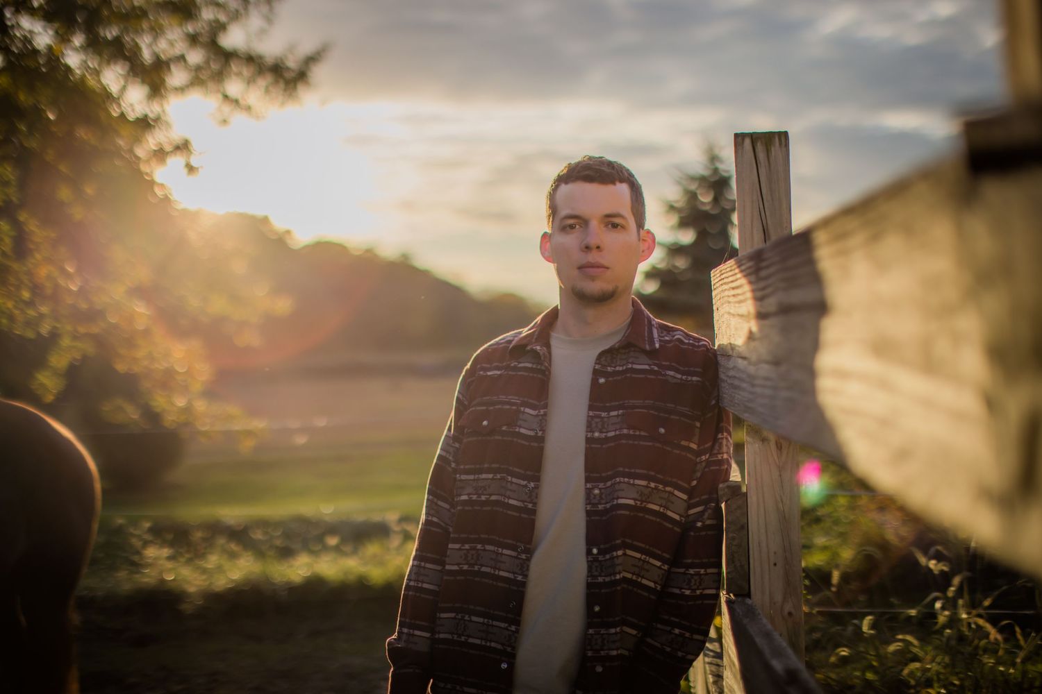 Man in striped flannel shirt poses against sunset in rural setting.