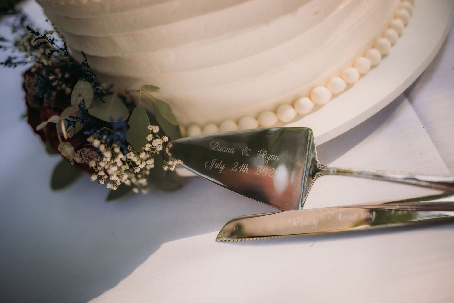 Elegant pearl-bordered wedding cake with silver serving utensils on white tablecloth.