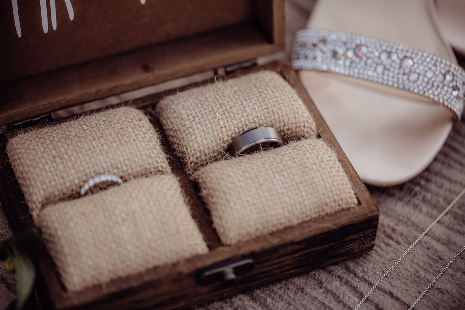 Wedding rings nestled in burlap-lined wooden box with crystal sandal visible.