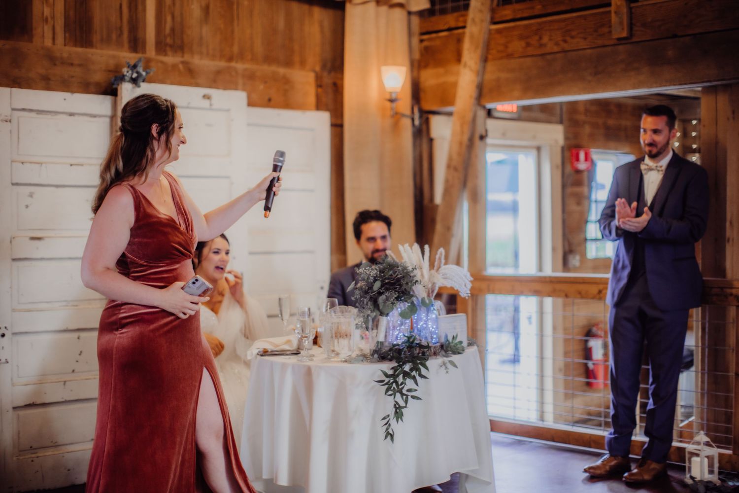 Wedding toast being given by speaker in burgundy dress at rustic barn reception.