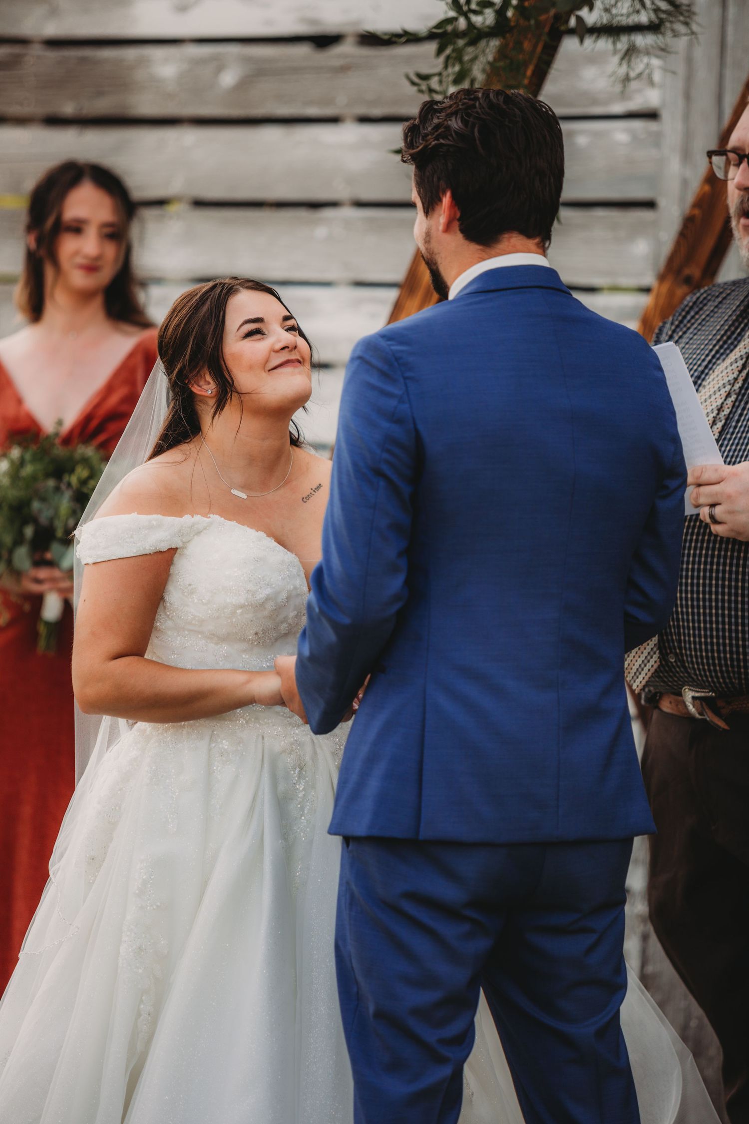 Wedding ceremony unfolds in front of a weathered barn wall with decorative wooden arch and greenery.