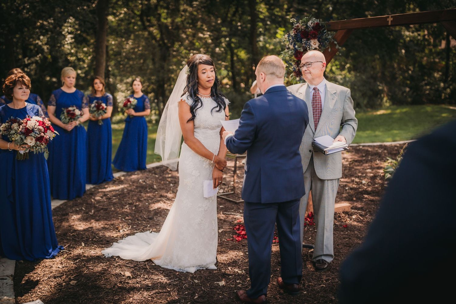 Wedding ceremony in progress at an outdoor wooded venue with blue-clad attendants.