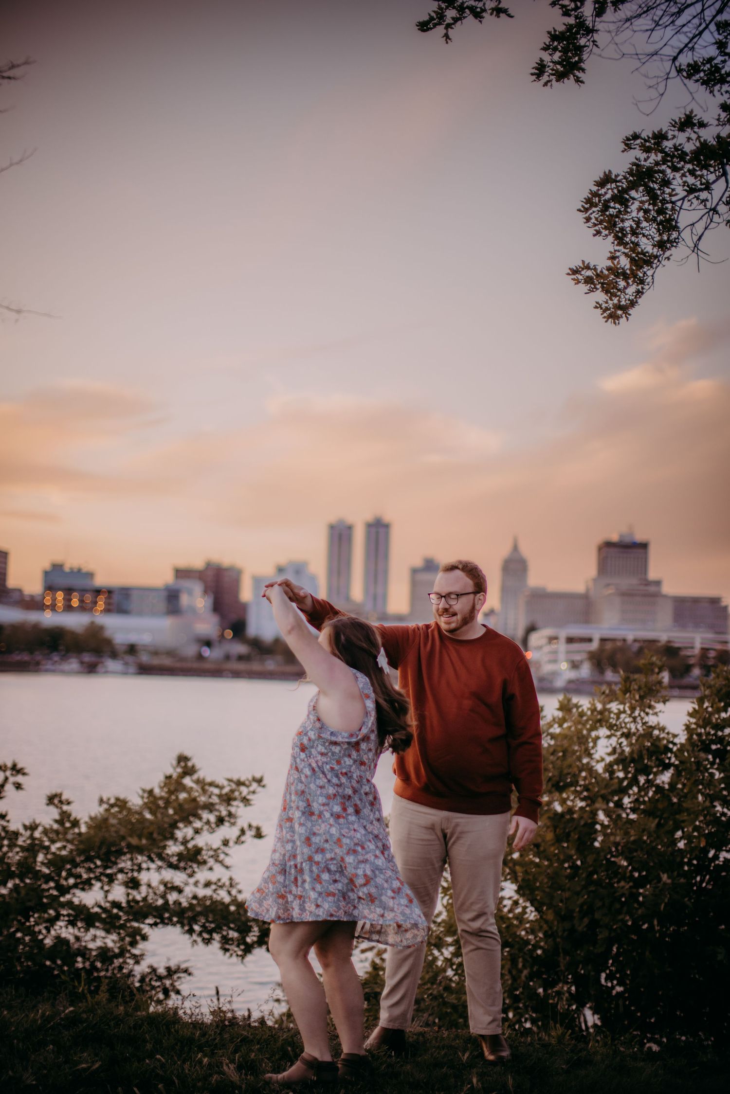 Couple dancing together on grassy hillside overlooking city skyline at dusk.