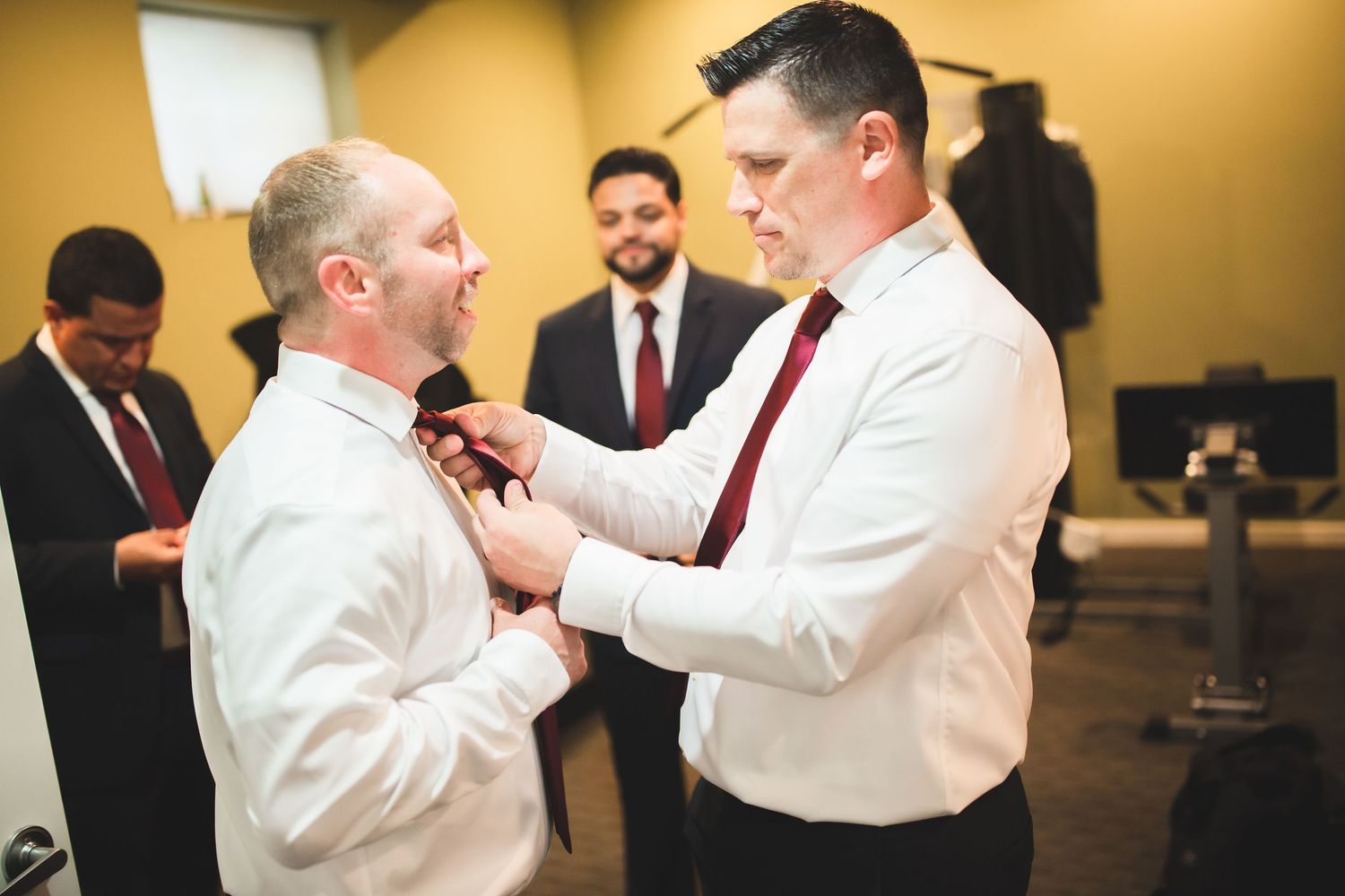 Two people in white shirts and red ties help each other with formal attire preparations.