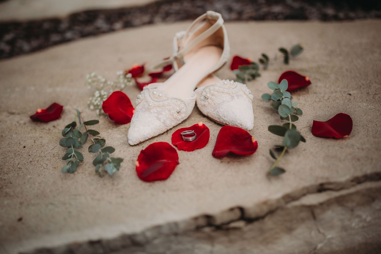 White lace wedding shoes with red rose petals and wedding rings arranged on stone surface.