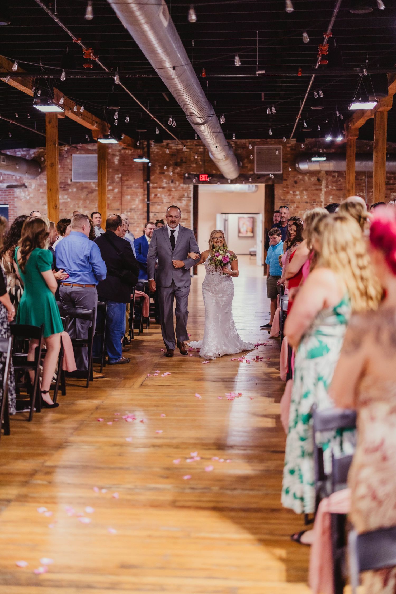 Wedding ceremony processional in a rustic brick venue with exposed pipes and wooden floors.