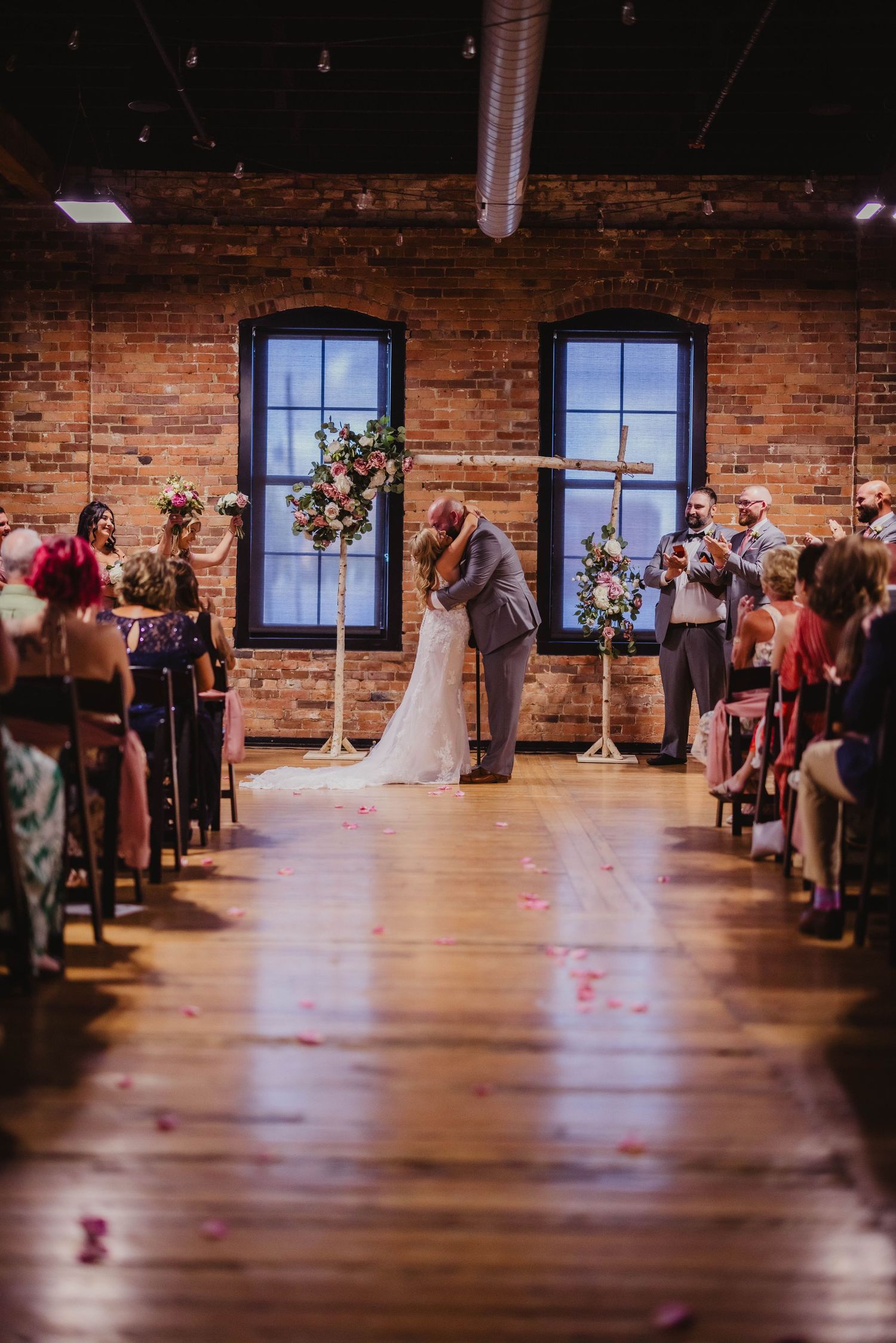 Couple shares intimate moment during wedding ceremony in rustic brick-walled venue with wooden floors and floral decor.