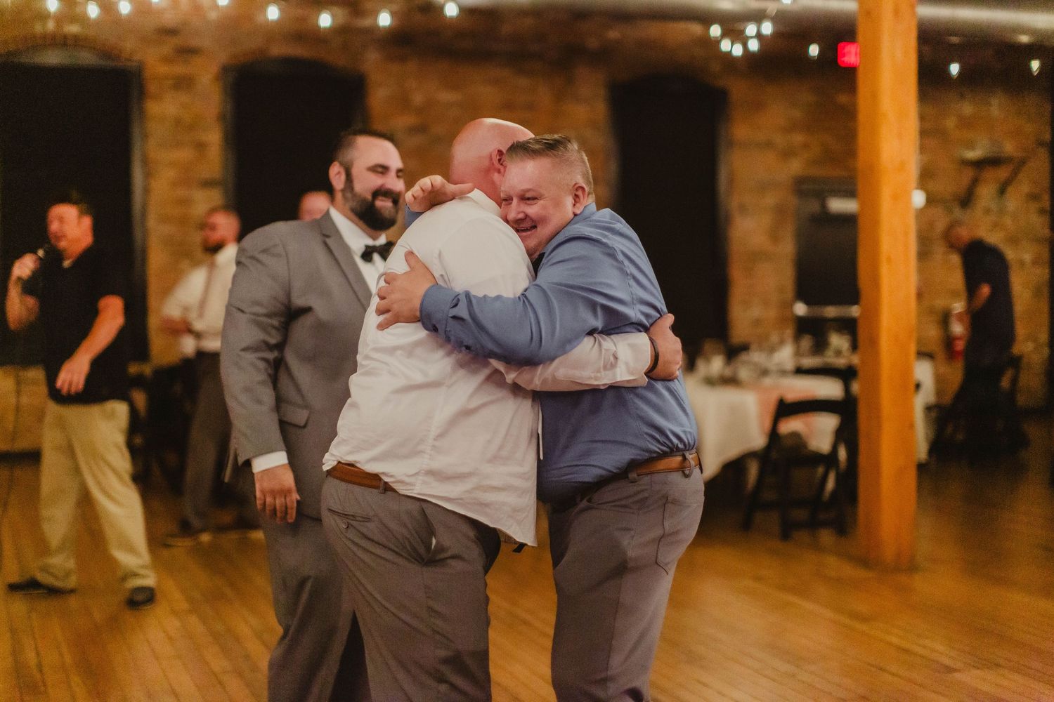 Wedding guests enjoy dancing in a rustic indoor venue decorated with string lights and wooden beams.