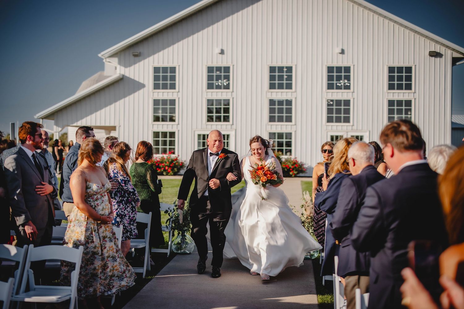 Wedding ceremony procession outside white barn venue with guests looking on.