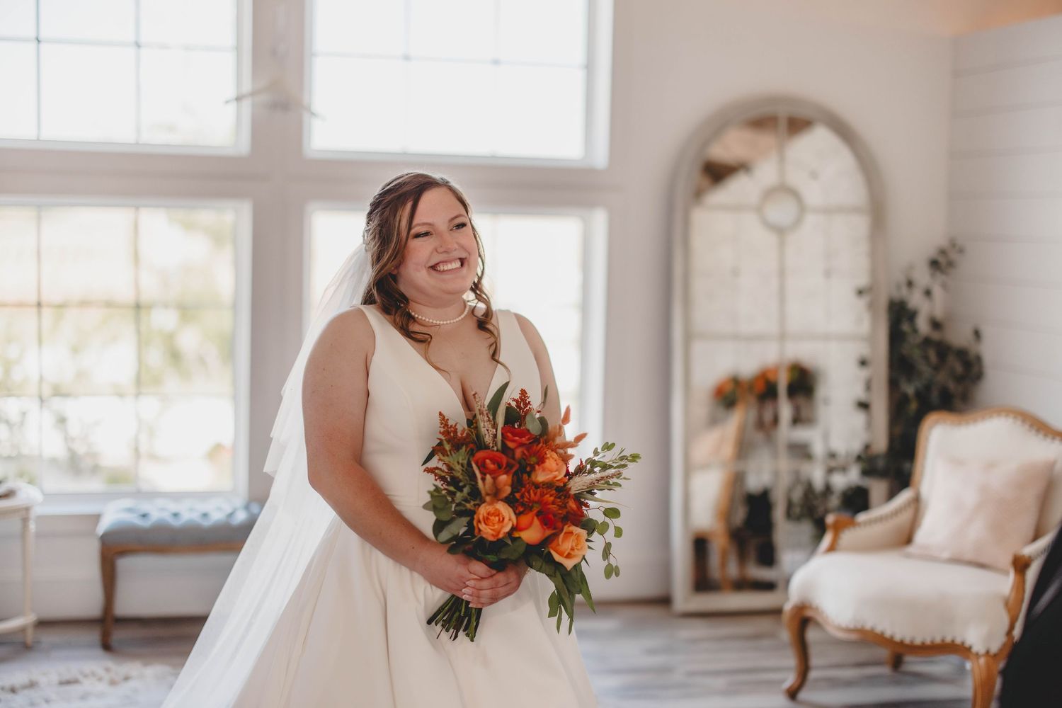 A bride holding vibrant orange and red flowers stands in a bright white room with elegant furnishings.
