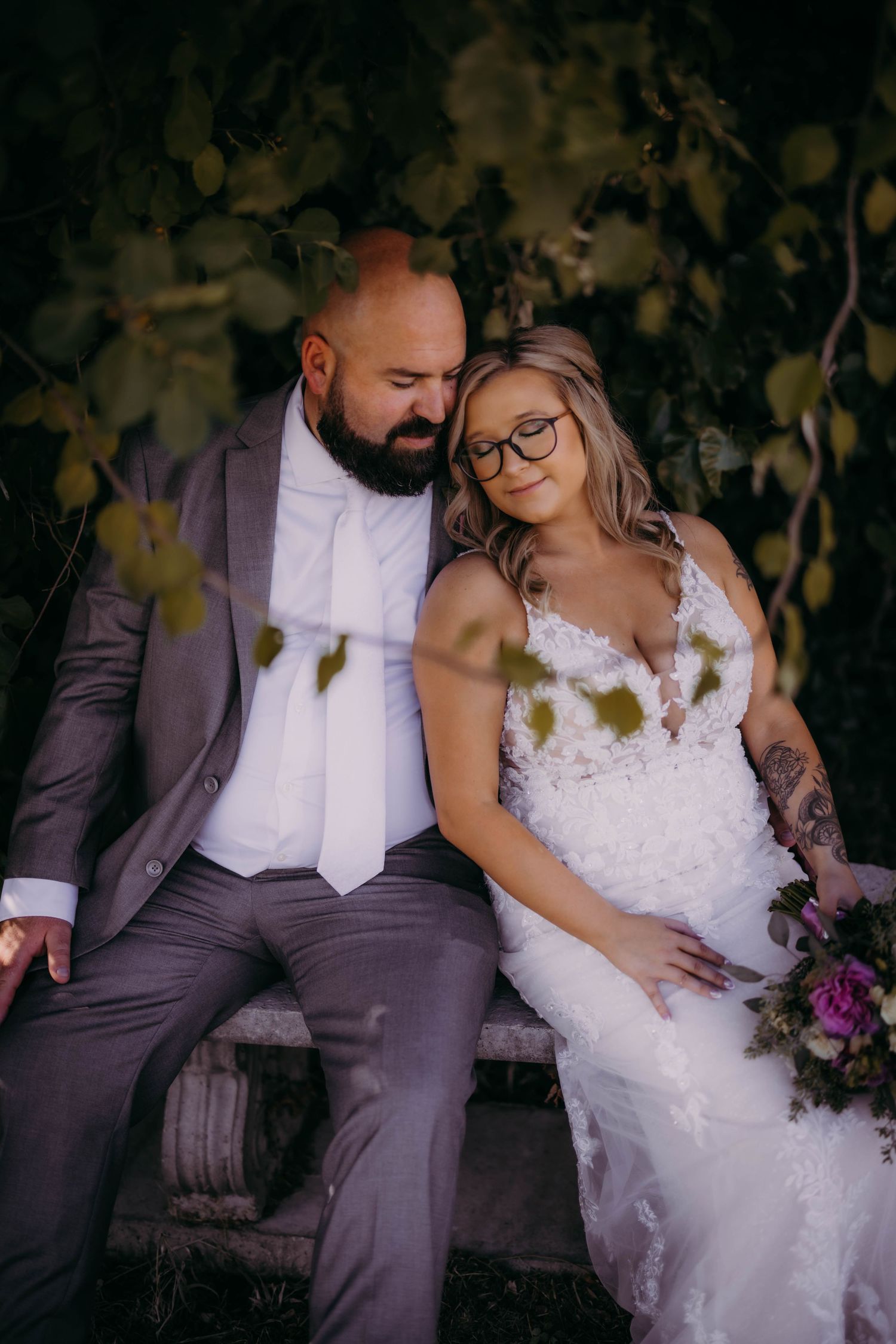 A tender moment between newlyweds sitting on a bench surrounded by greenery.