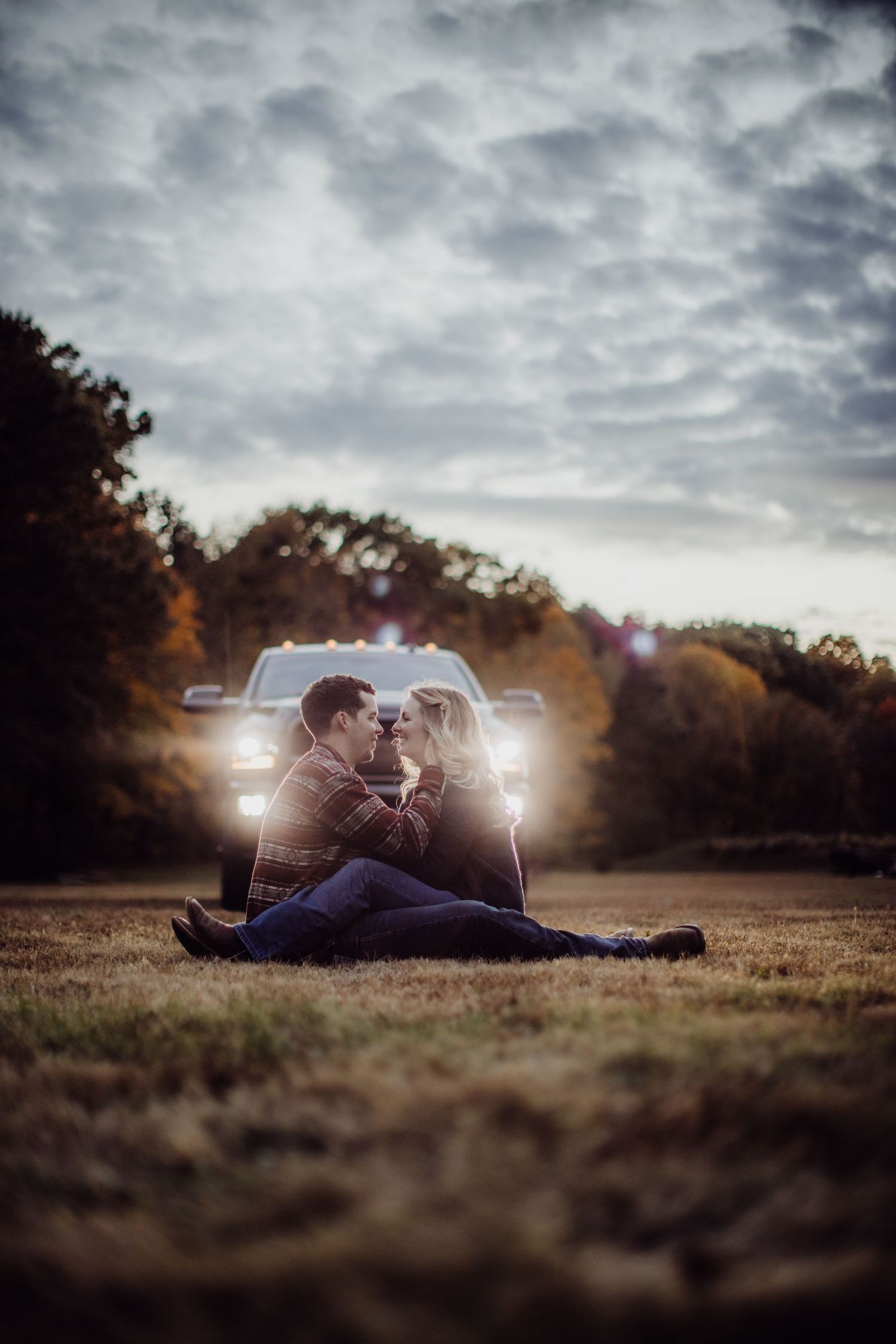 A romantic couple sits in a field at sunset with car headlights illuminating them from behind creating a dreamy atmosphere.