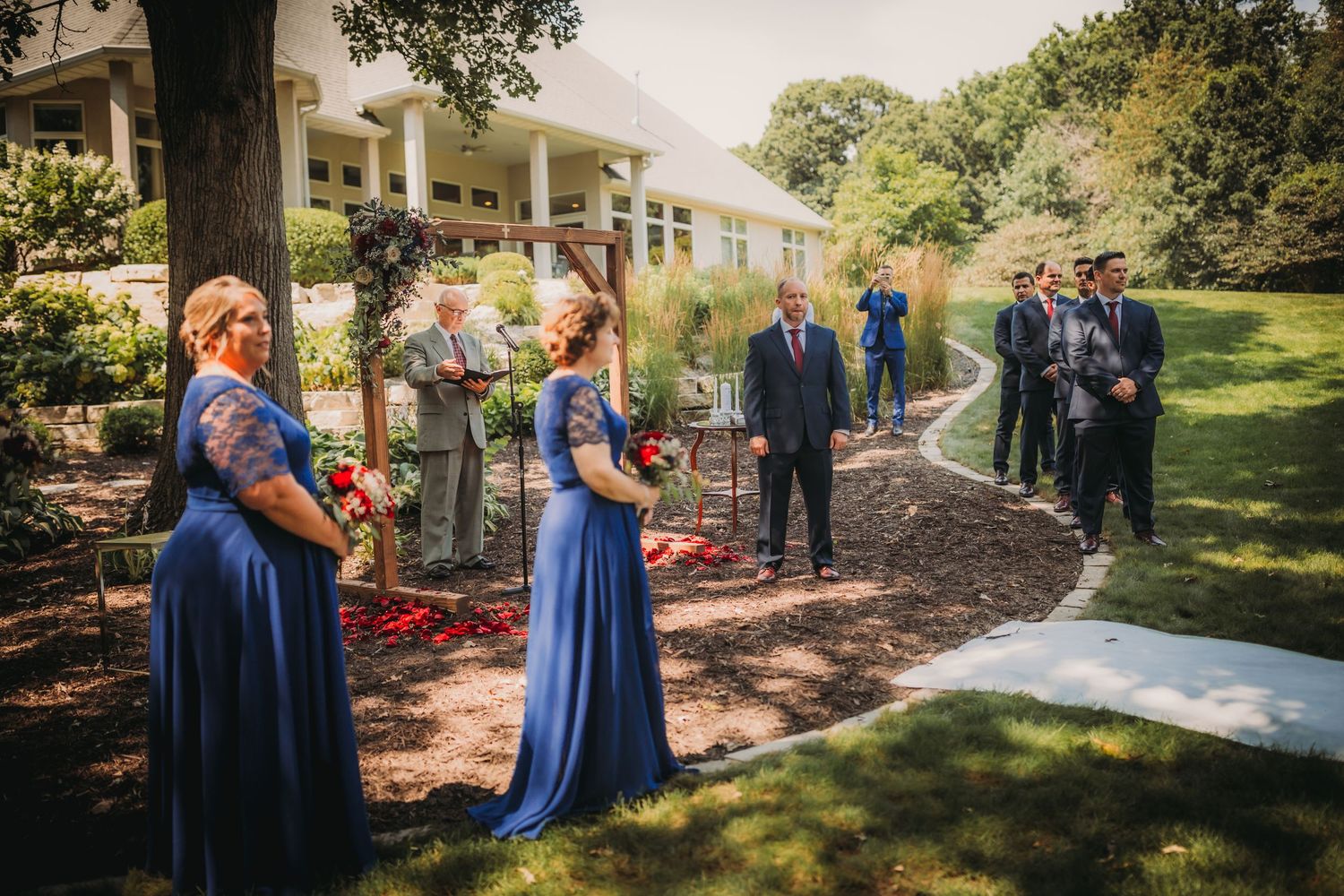 Wedding party in blue attire gathers along garden path for outdoor ceremony.