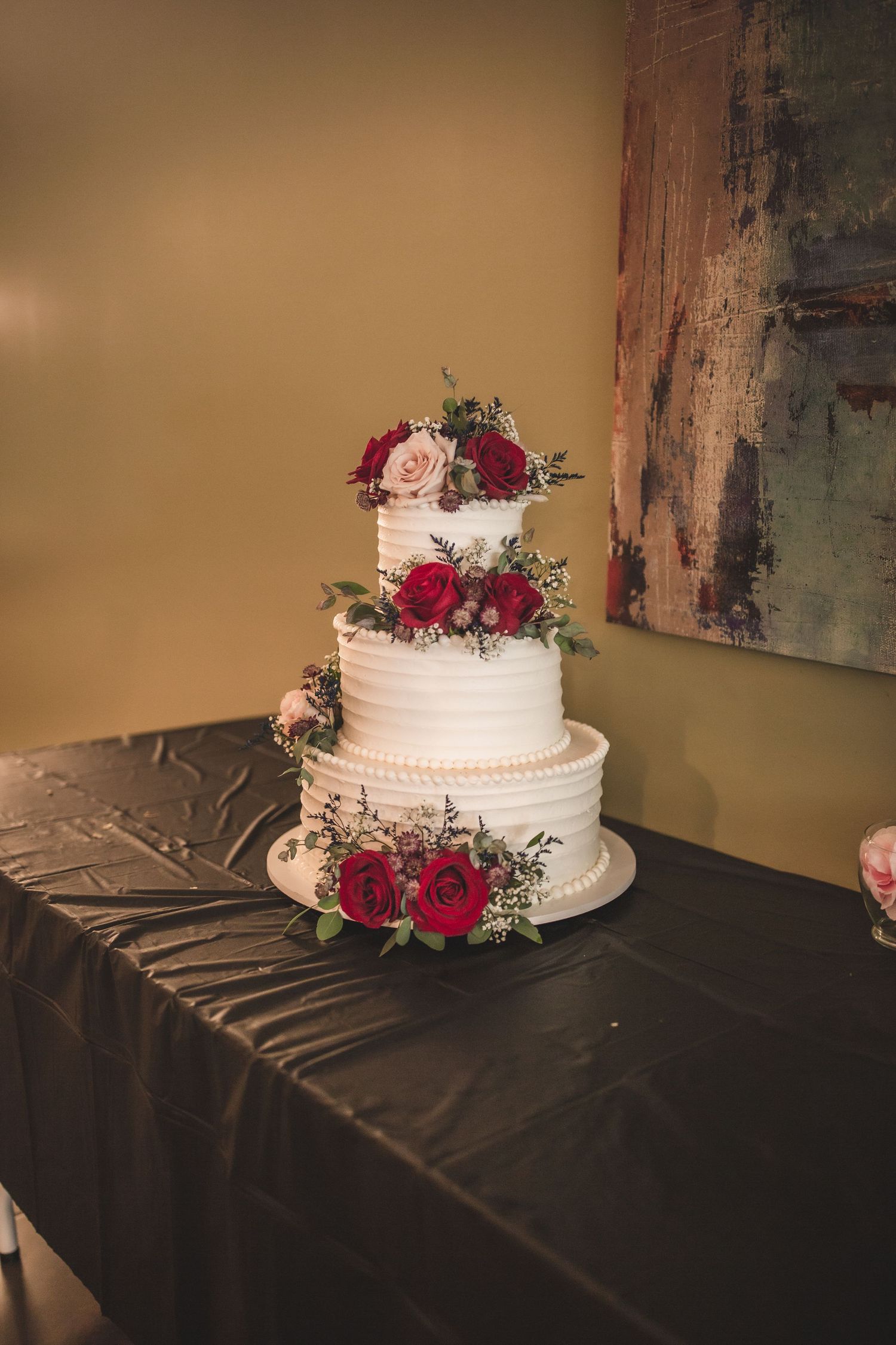 Elegant white wedding cake with red rose decorations displayed on black tablecloth.