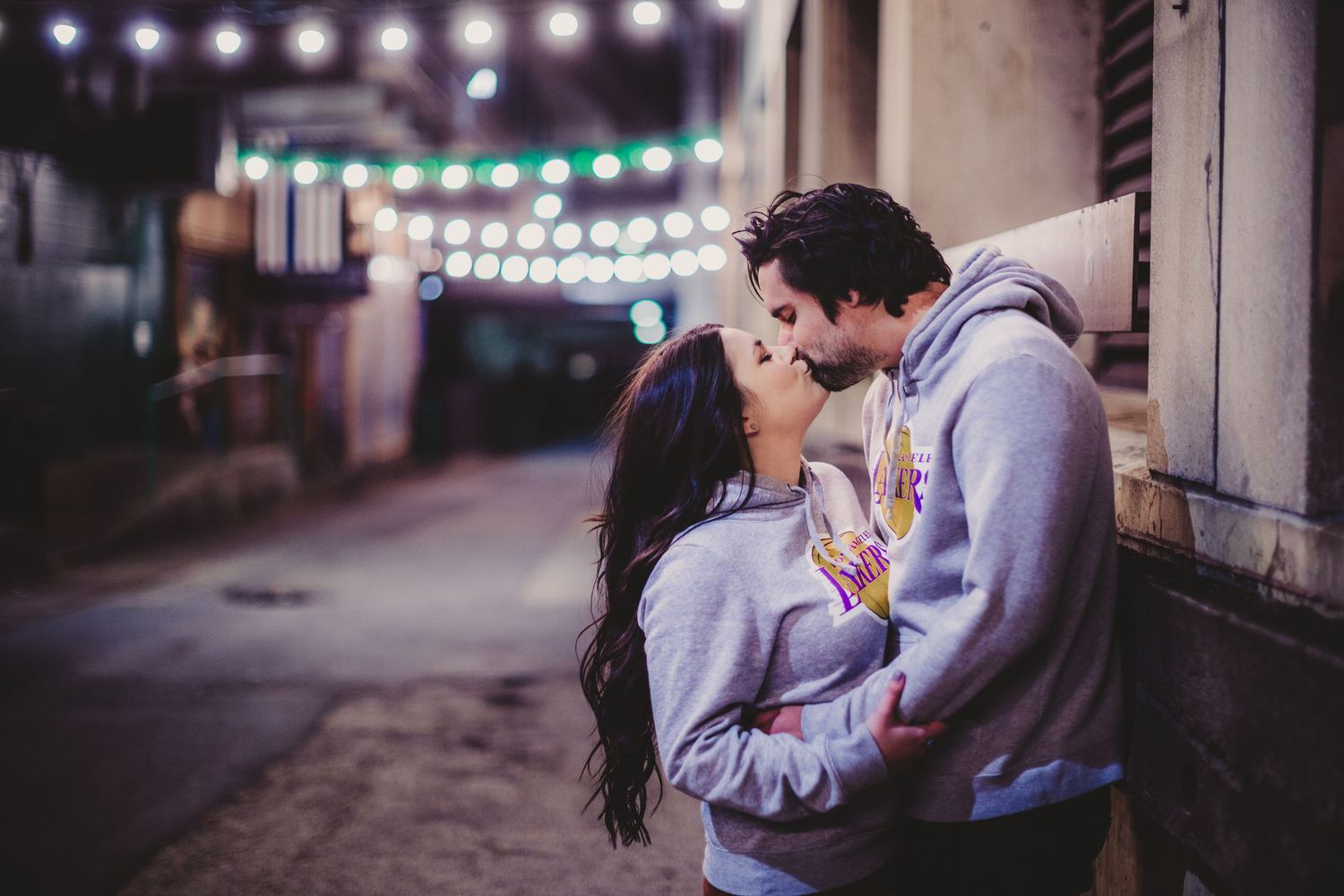 Romantic couple in matching gray hoodies kissing under decorative string lights at night.