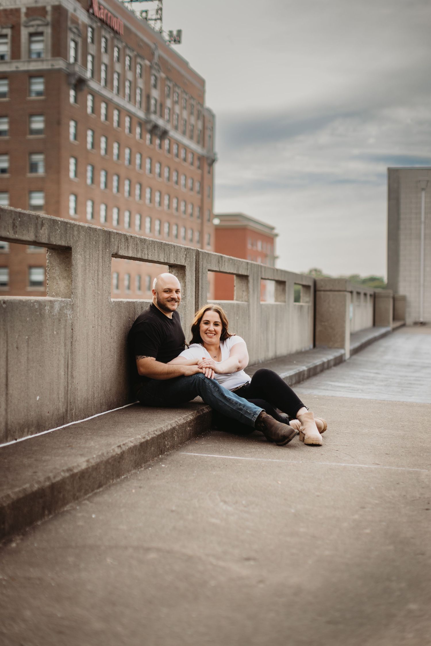 A couple relaxes together on a concrete barrier with brick buildings and cloudy skies in the background.