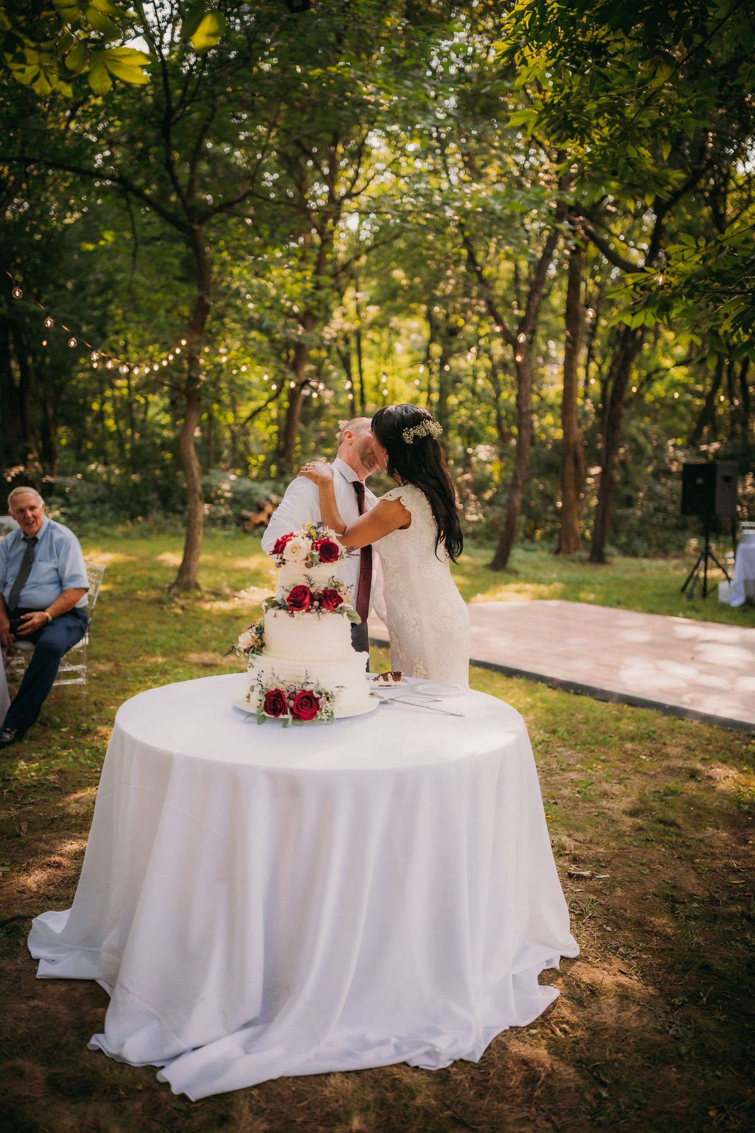 A couple cuts their three-tiered white wedding cake decorated with red flowers at a woodland ceremony.