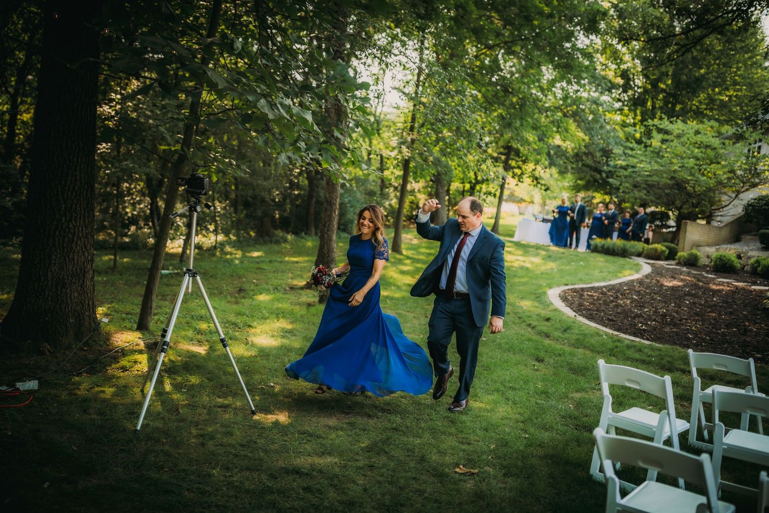 Wedding attendants walk down an outdoor garden path with white chairs and lush greenery.