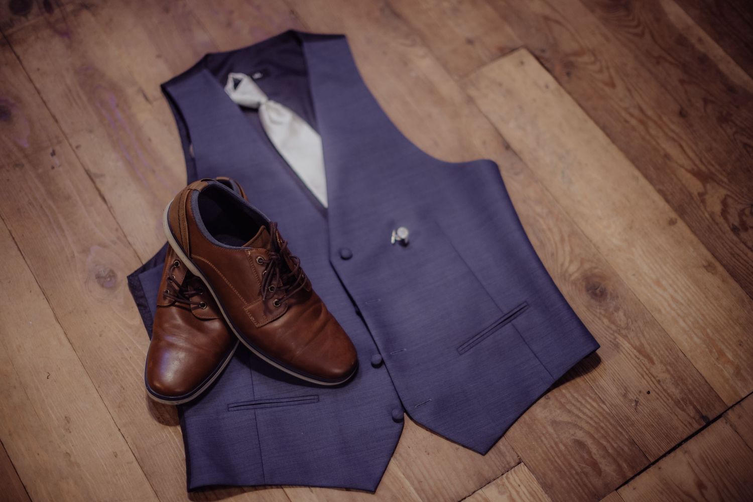 Navy blue vest and brown leather dress shoes arranged on wooden floor.