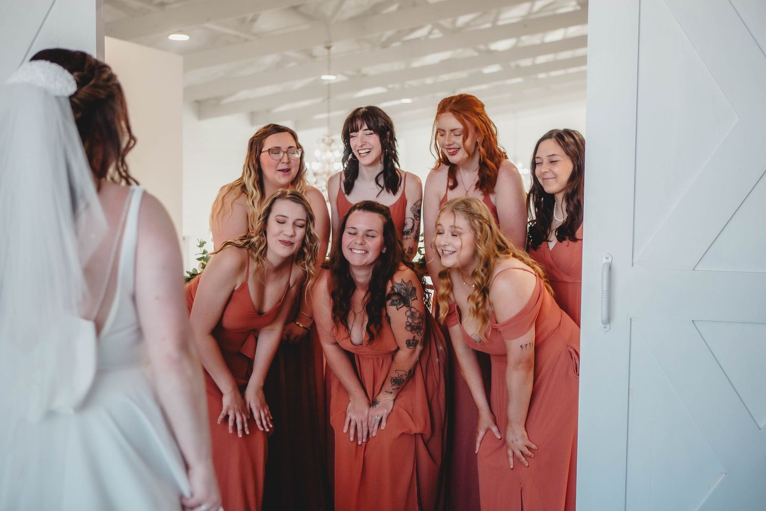Bridesmaids in coral dresses react with joy upon seeing bride in her wedding dress.