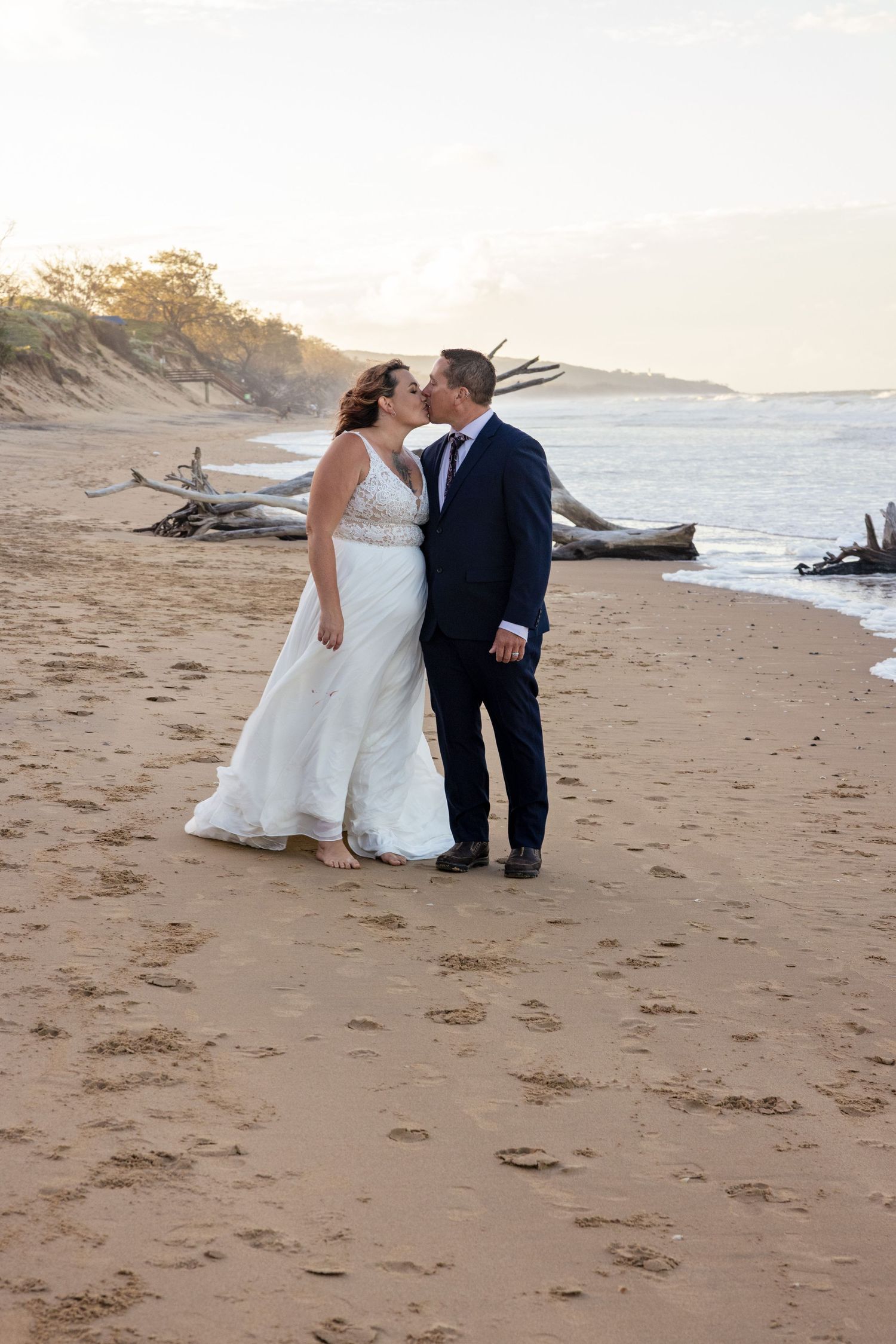 A couple in wedding attire share an intimate moment on a sandy beach at sunset with driftwood adding natural decor.